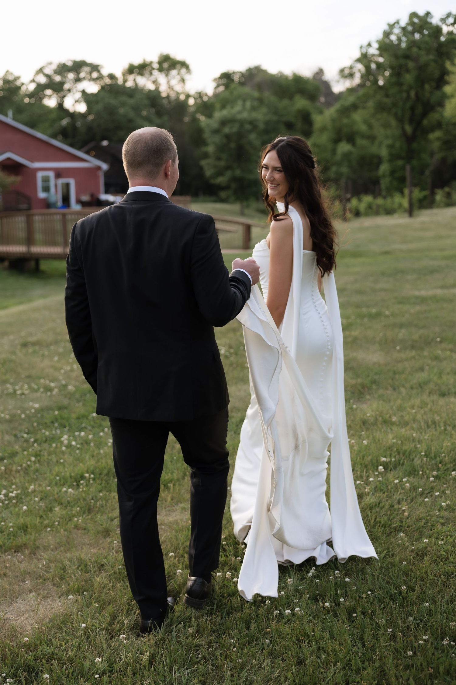Smiling bride in a white backless gown looks back at groom while walking hand in hand across a green lawn.