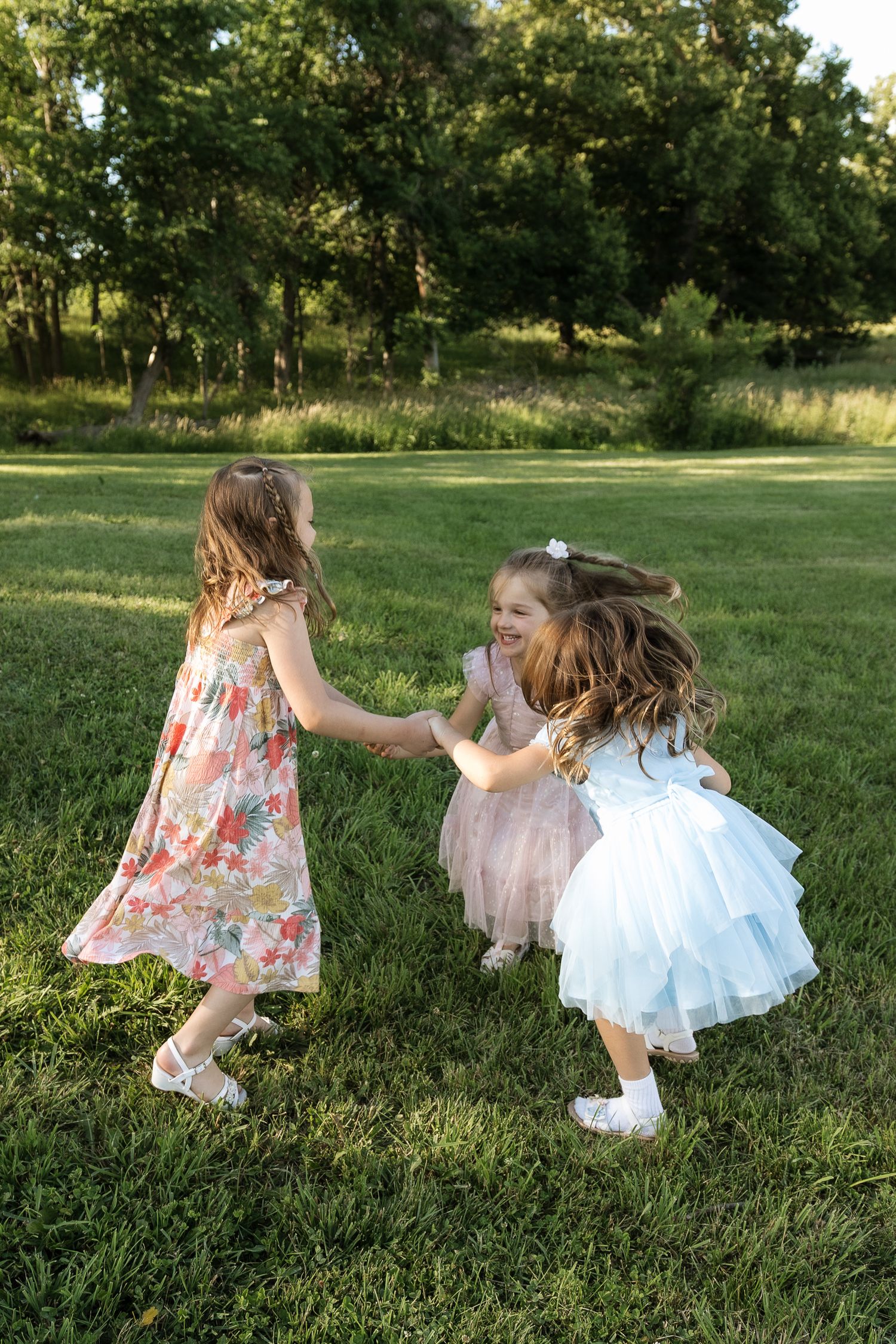 Three little girls in pretty dresses hold hands and laugh together in a park.