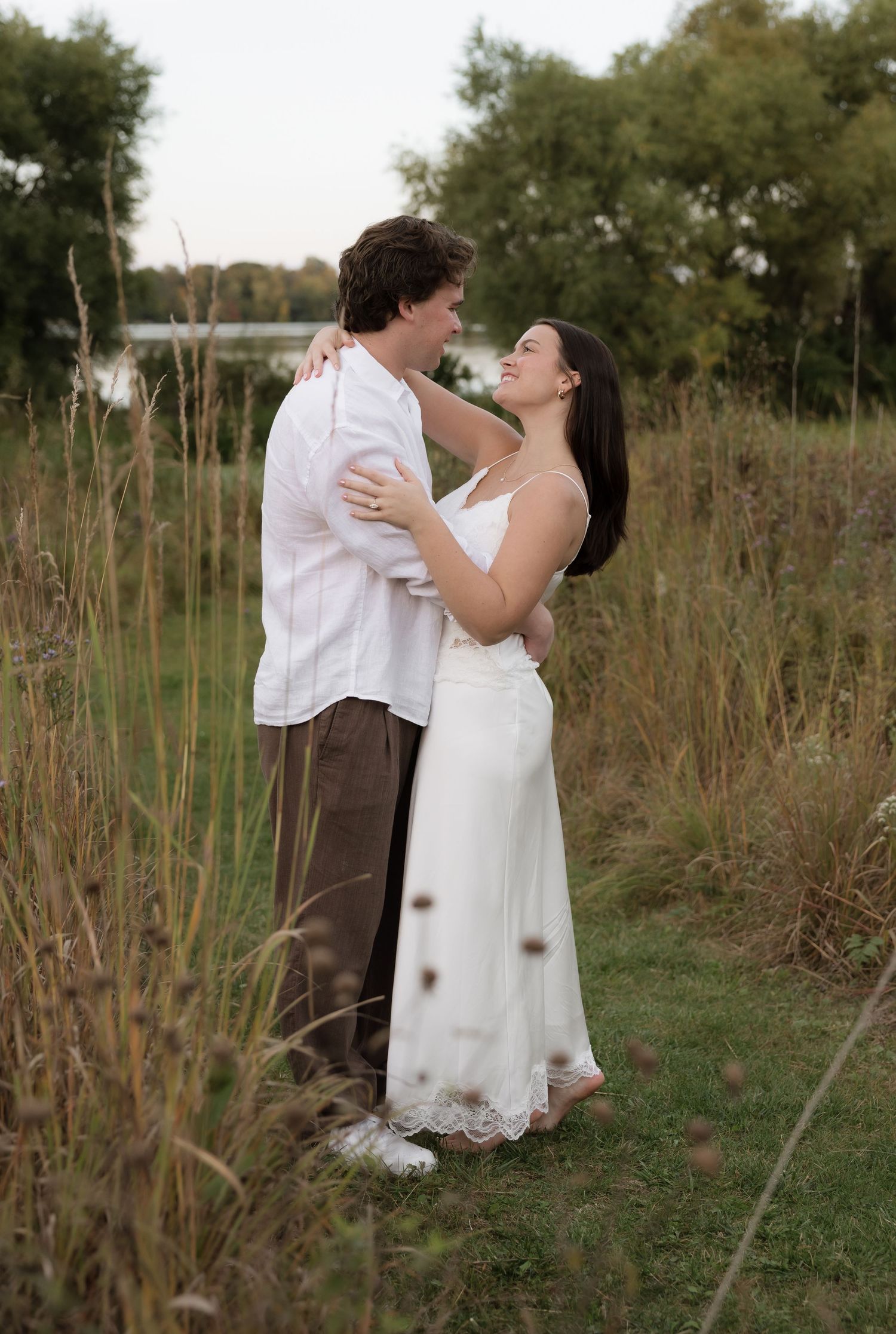 Romantic couple embracing in a field of tall grass, woman in white dress, man in white shirt, near a lake.