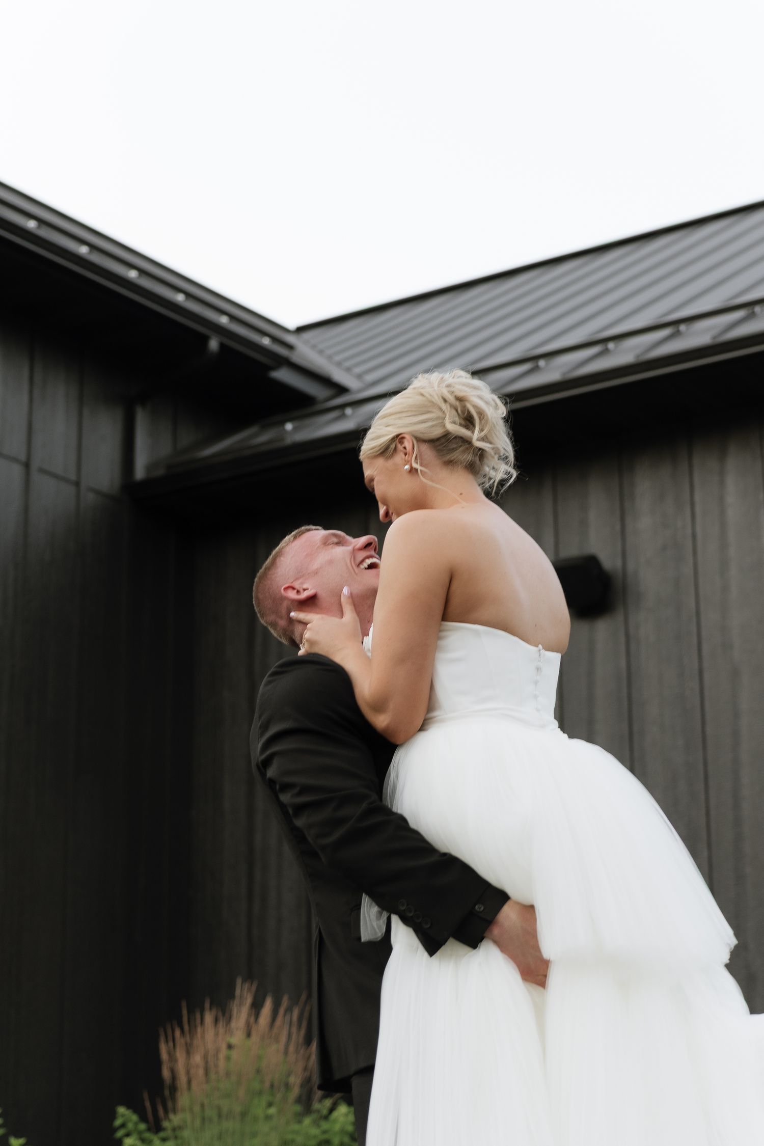 Groom lifts laughing bride in white gown outside dark barn wedding venue.