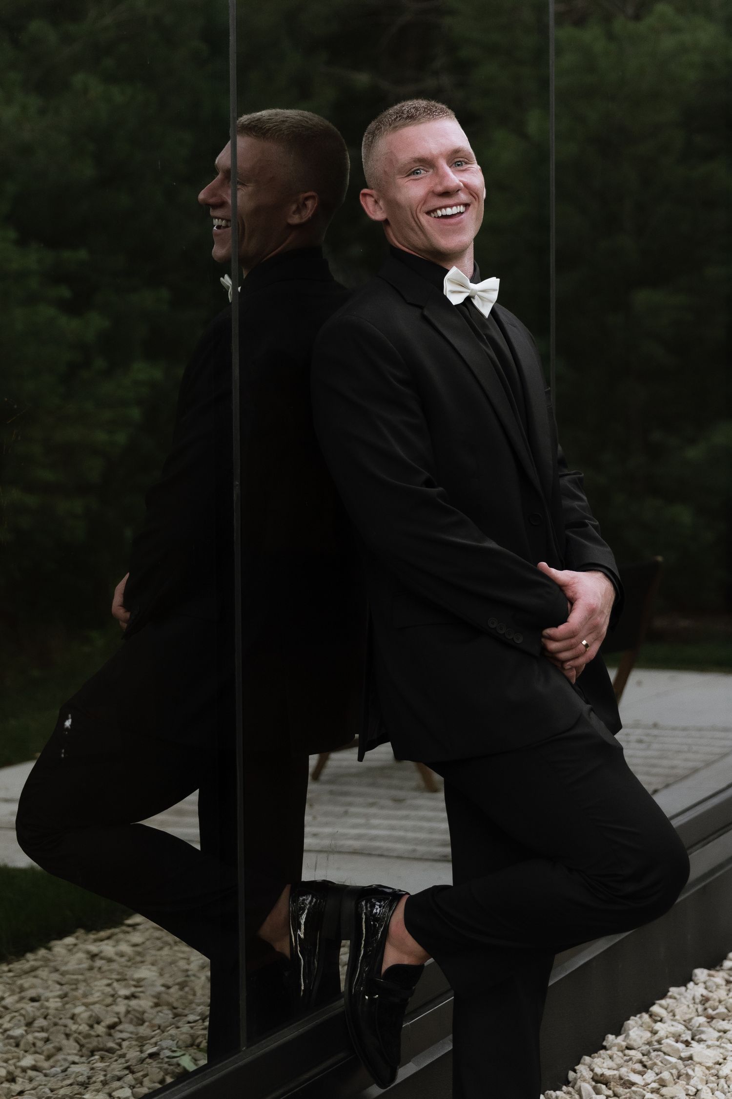 Smiling groom in black tuxedo with white bow tie leaning against mirrored wall with reflection visible.