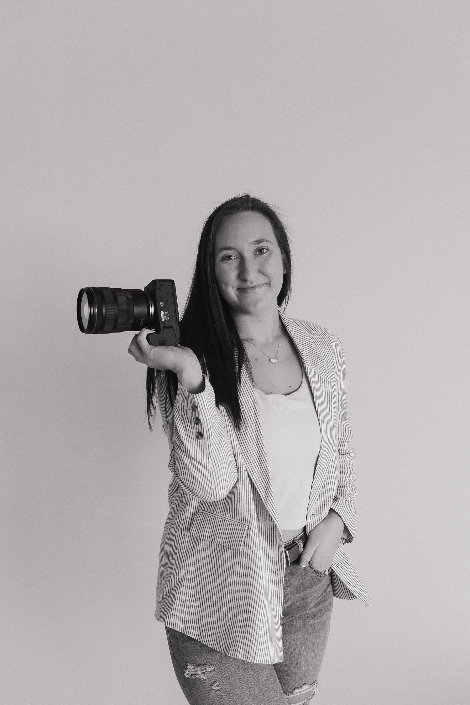 Black and white portrait of female photographer holding DSLR camera wearing blazer and ripped jeans.