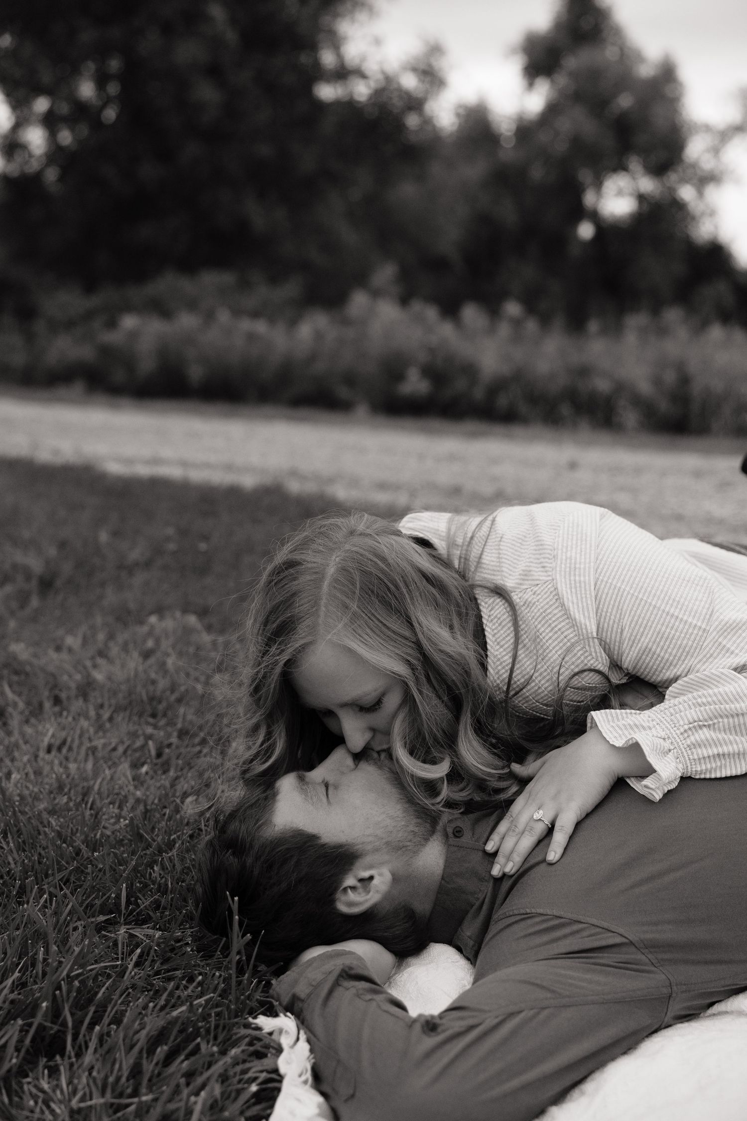 Black and white photo of a couple sharing a romantic kiss outdoors, woman leaning over man lying on grass.