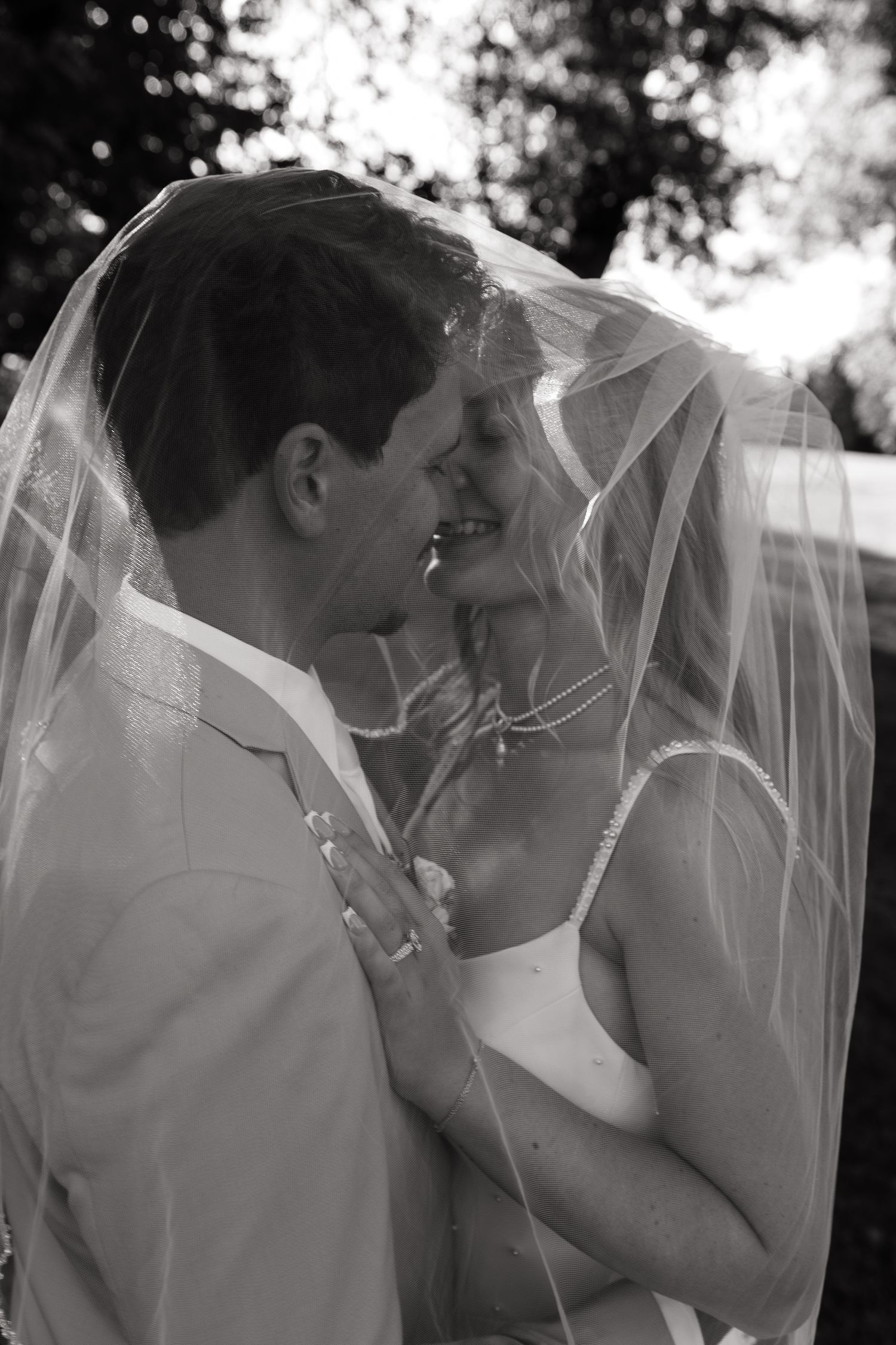 Black and white photo of a bride and groom sharing a romantic moment under the wedding veil outdoors.