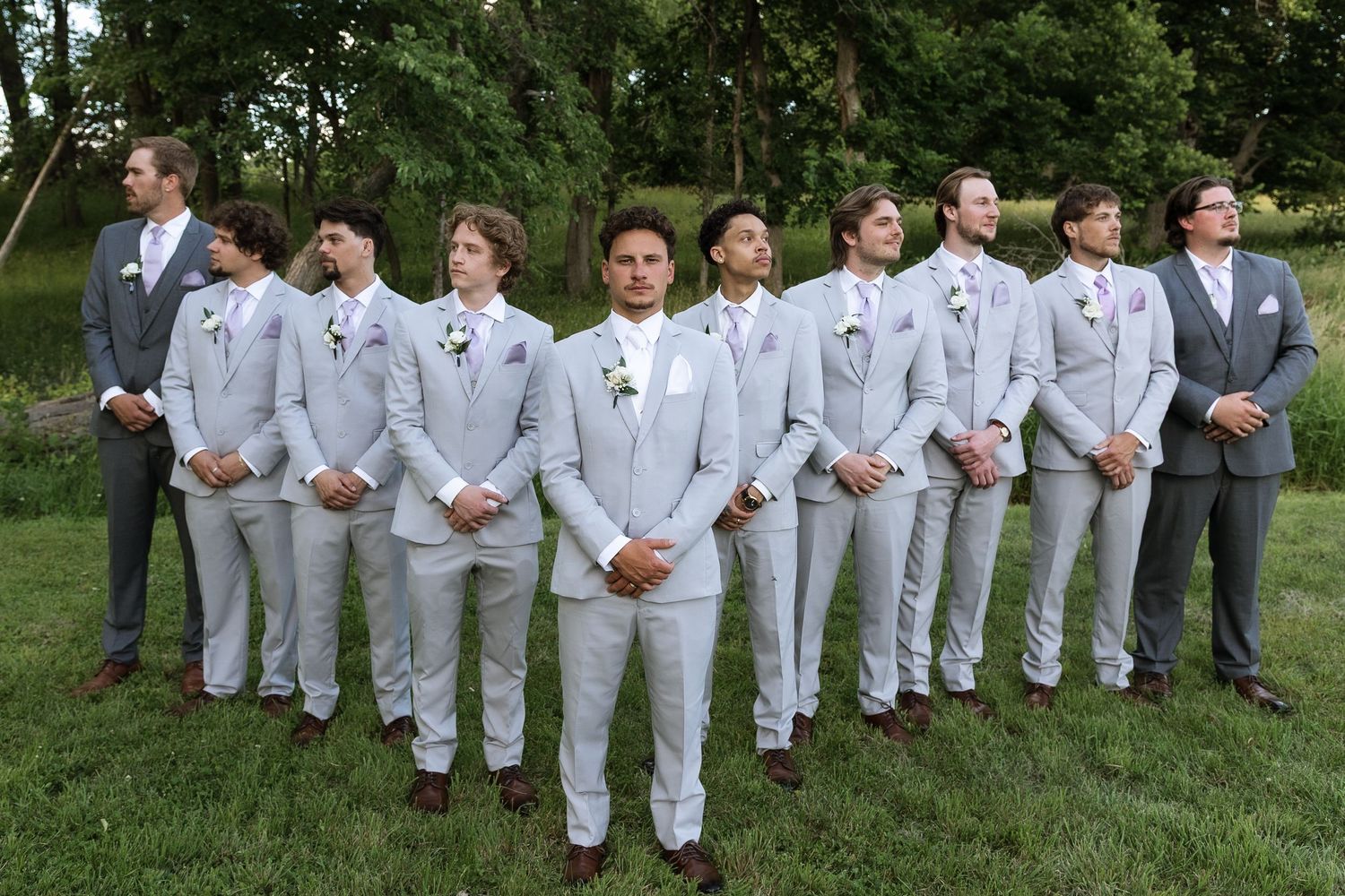 Groom and groomsmen in light gray suits posing outdoors on green grass for a wedding party photo.