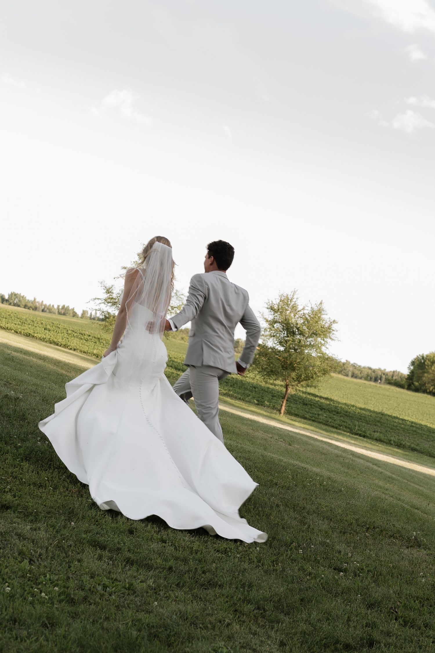 Bride in white gown and veil walks with groom in gray suit across lush green lawn at outdoor wedding venue.