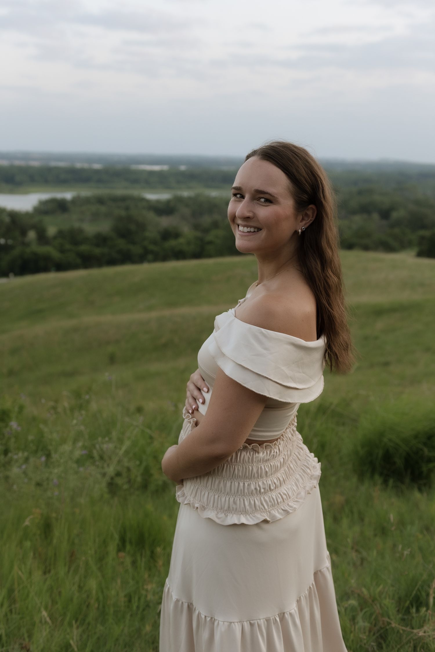Smiling young woman in off-shoulder cream dress poses on green hillside with scenic lake view backdrop.