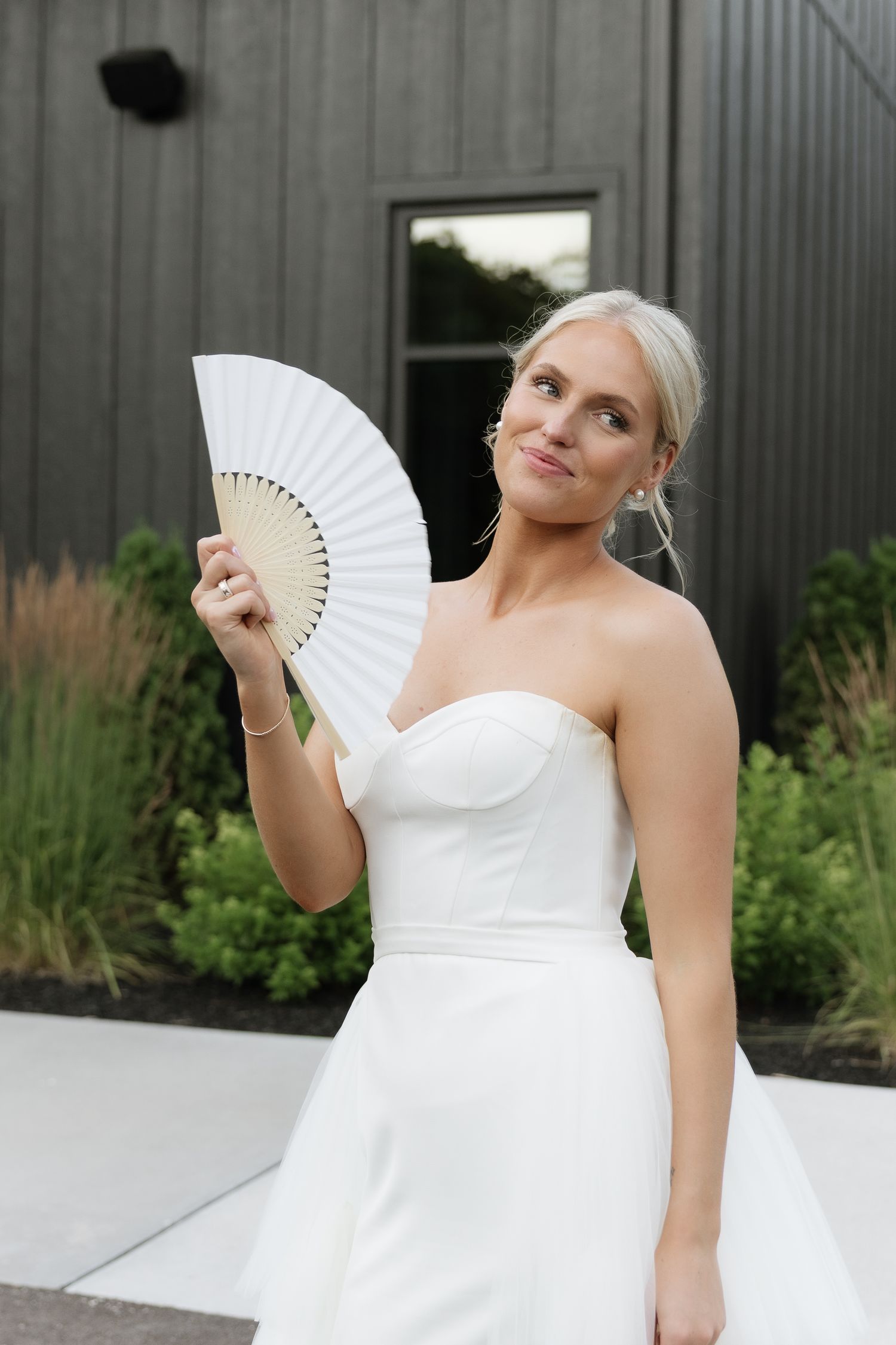 Blonde bride in strapless white gown holding a white paper fan outdoors near a dark barn venue.