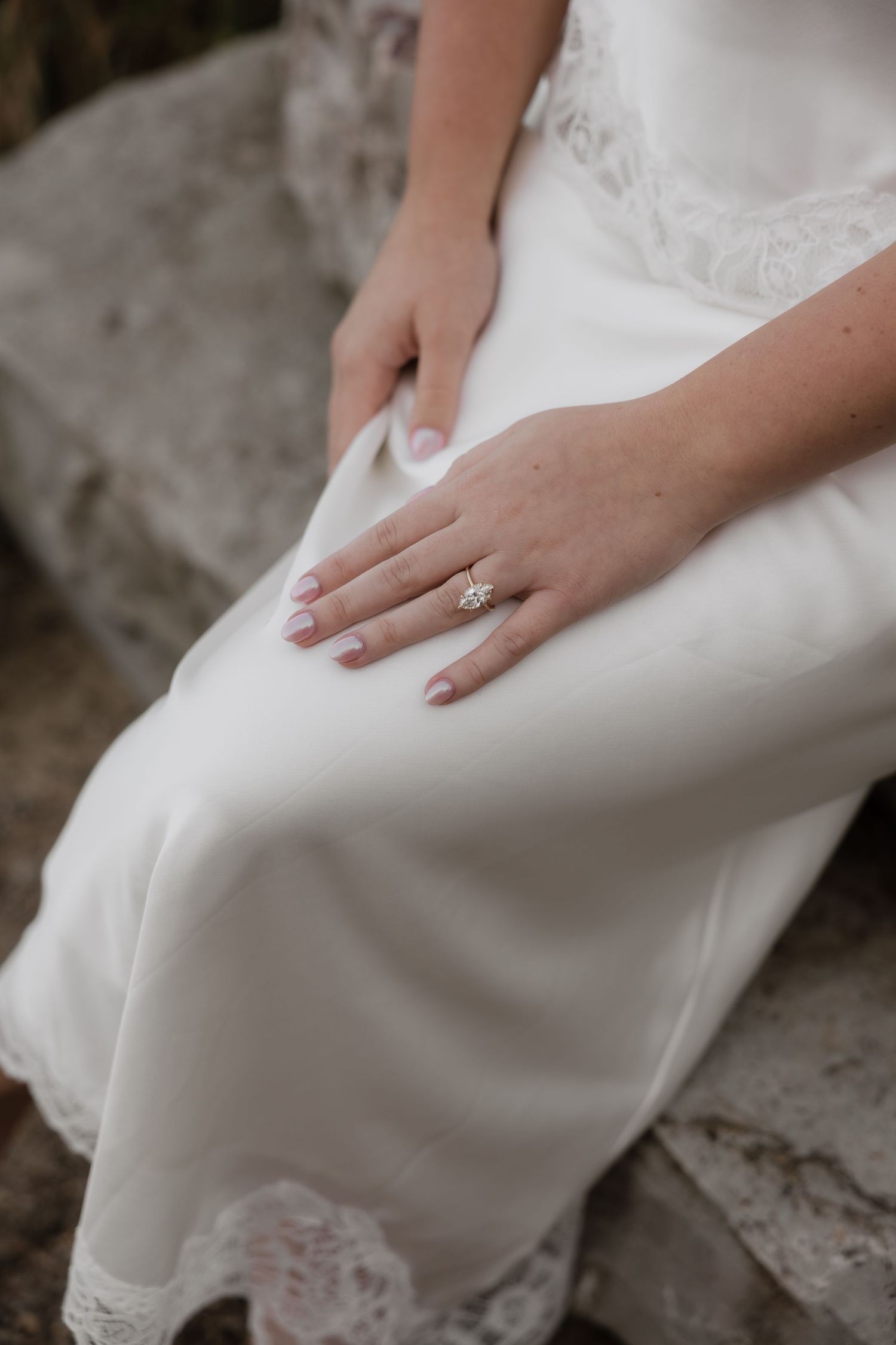 Close-up of bride's hand displaying a stunning diamond engagement ring on white satin lace dress.