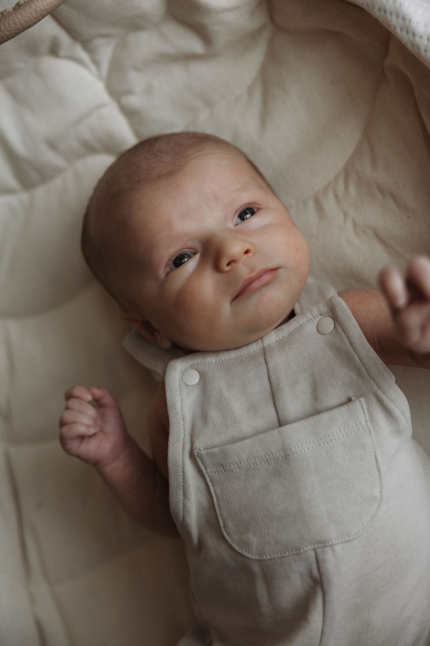 Newborn baby in beige linen romper lying in cream bassinet looking upward.