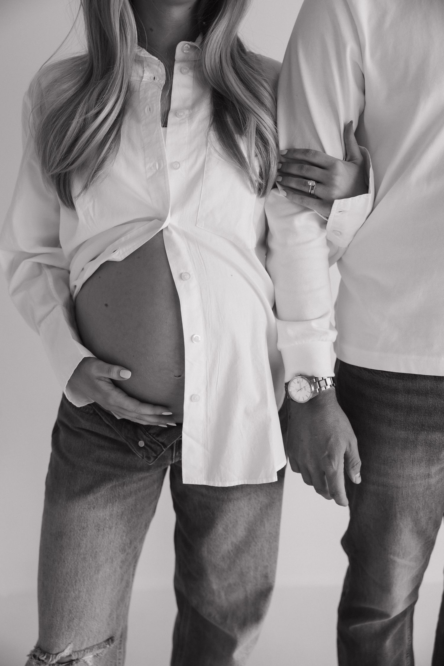 Black and white maternity photo of pregnant woman in white shirt and jeans with partner holding her bump.