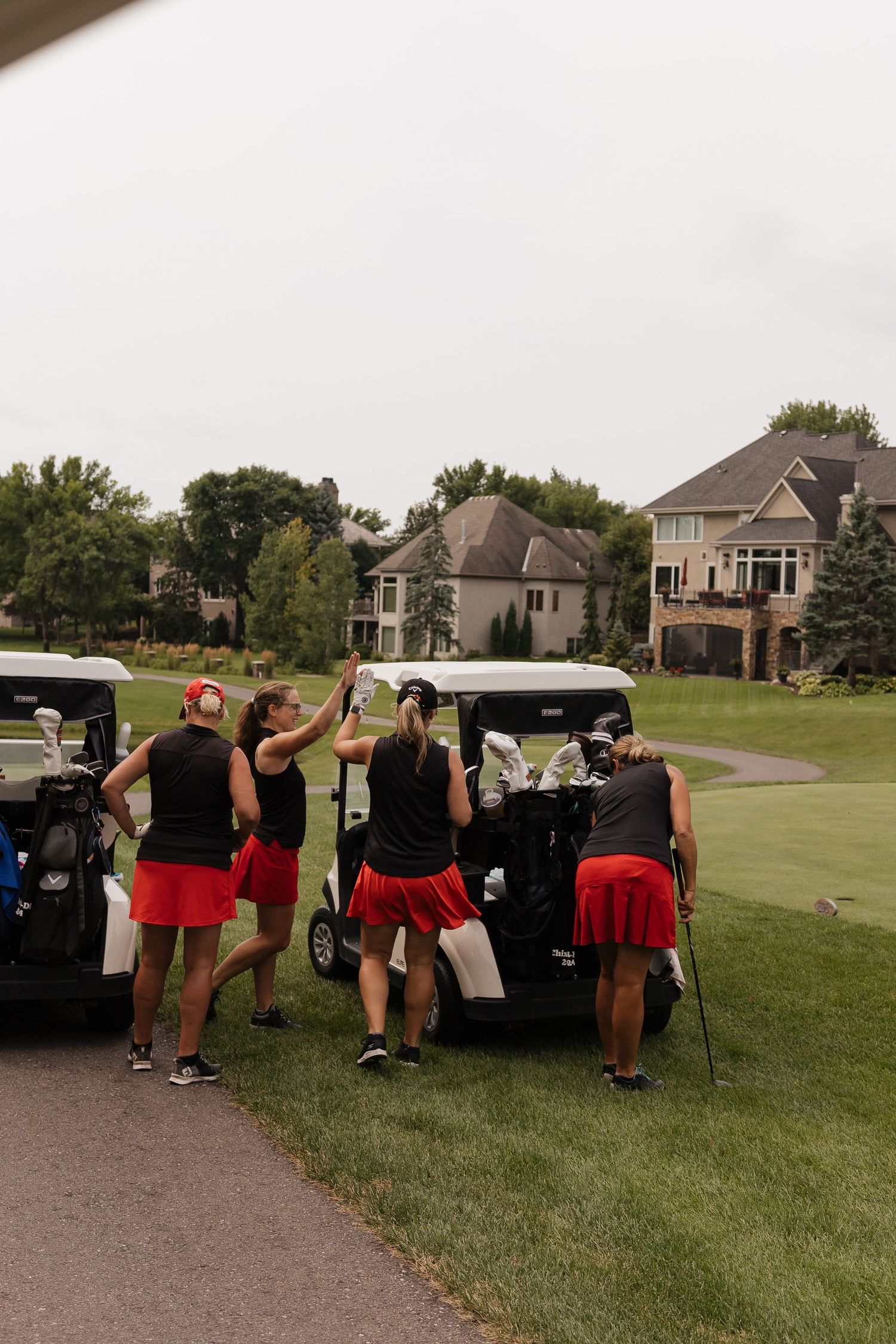Women in red skirts and black tops gather around a golf cart on a lush golf course near upscale homes.