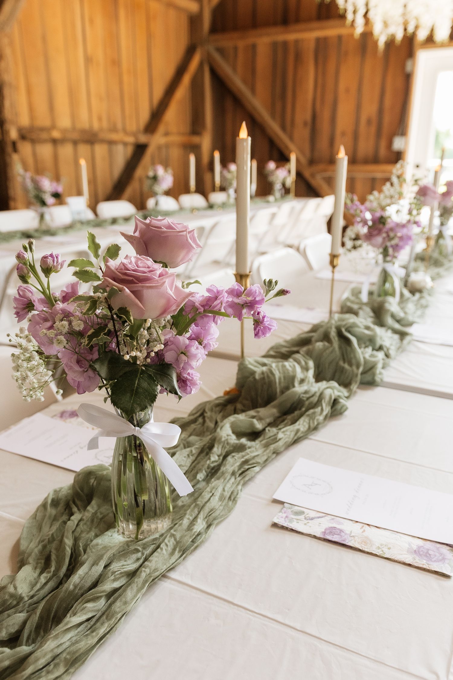 Elegant wedding reception table with purple roses, candles, and sage green gauze runner in rustic barn venue.