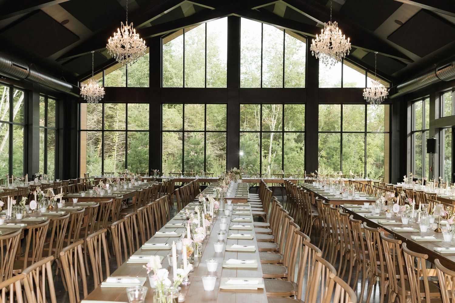 Elegant wedding reception hall with long dining tables, wooden chairs, chandeliers, and floor-to-ceiling windows overlooking lush greenery.