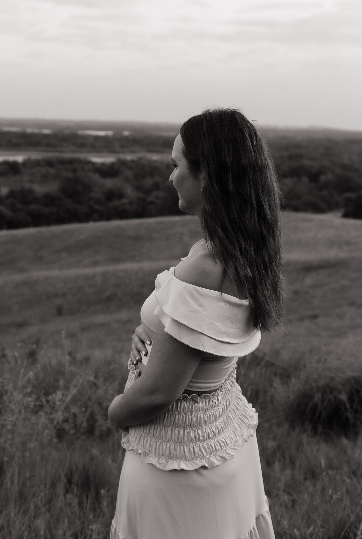 Black and white photo of a young woman in an off-shoulder dress standing in a field, gazing into the distance.