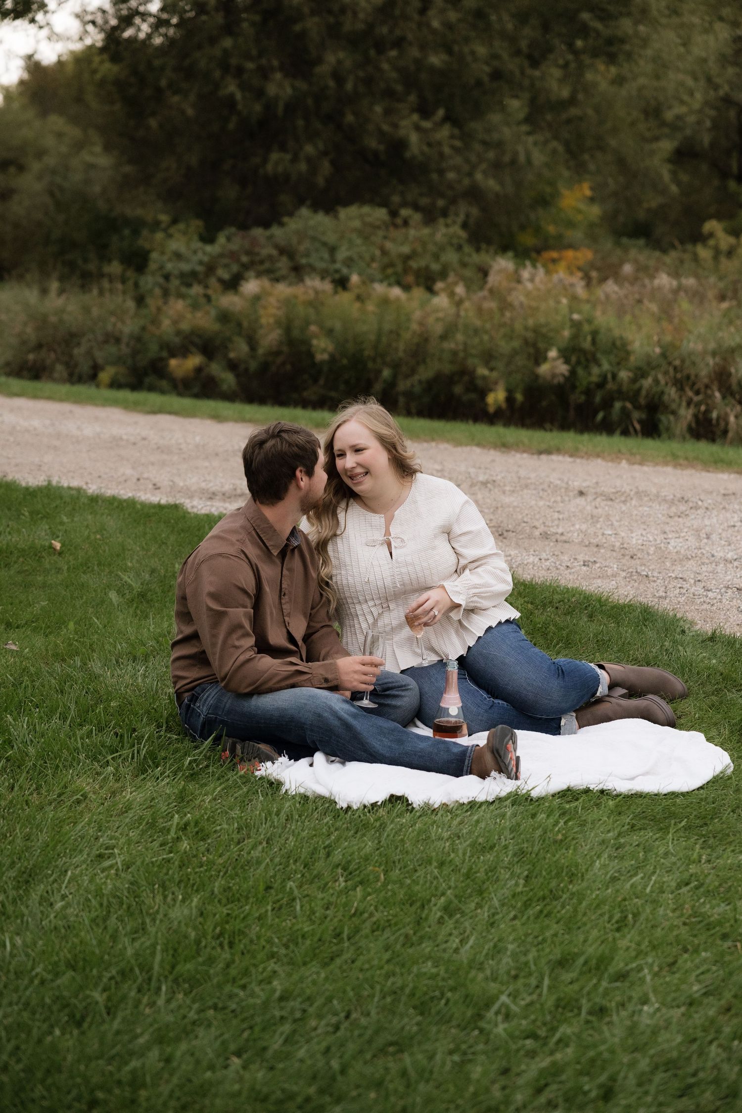 A couple sits on a white blanket in a park, laughing together during an autumn outdoor engagement photo session.
