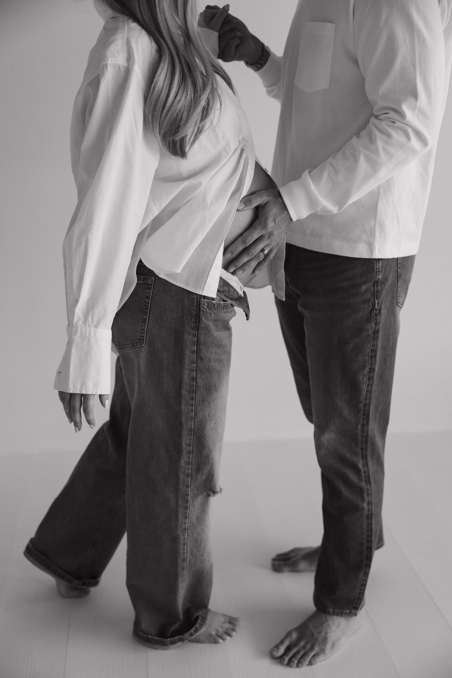 Black and white image of couple in white shirts and jeans, man placing hand on pregnant partner's baby bump.