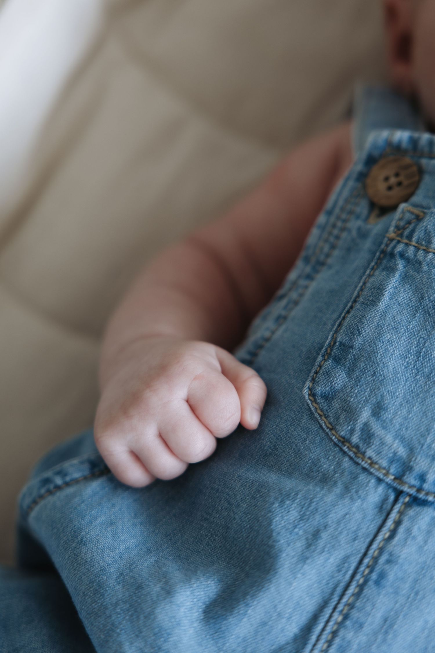 Close-up of newborn baby's tiny fist gripping blue denim overalls pocket.