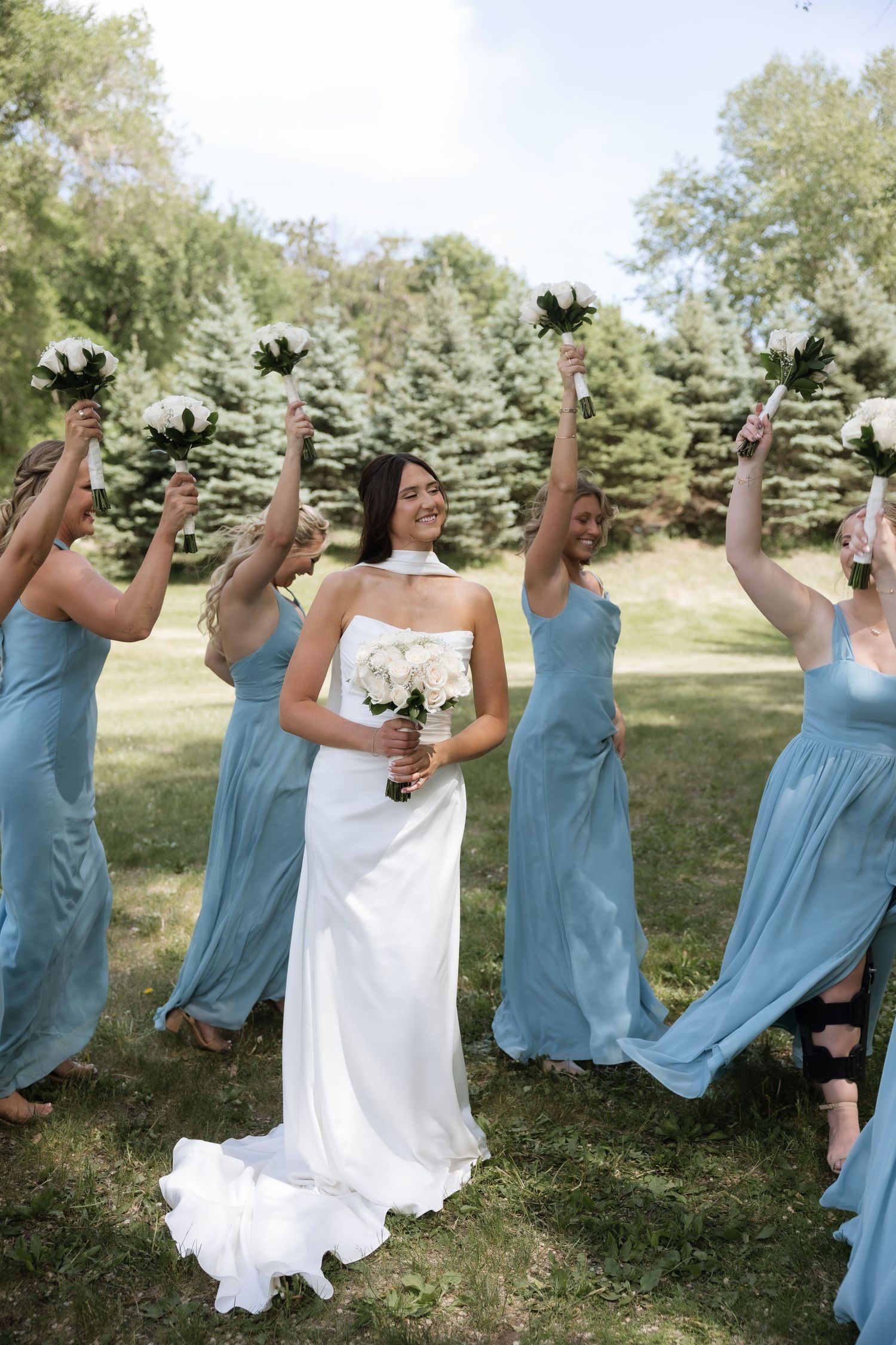 Bride in a white halter gown smiles as bridesmaids in dusty blue dresses raise bouquets in celebration.