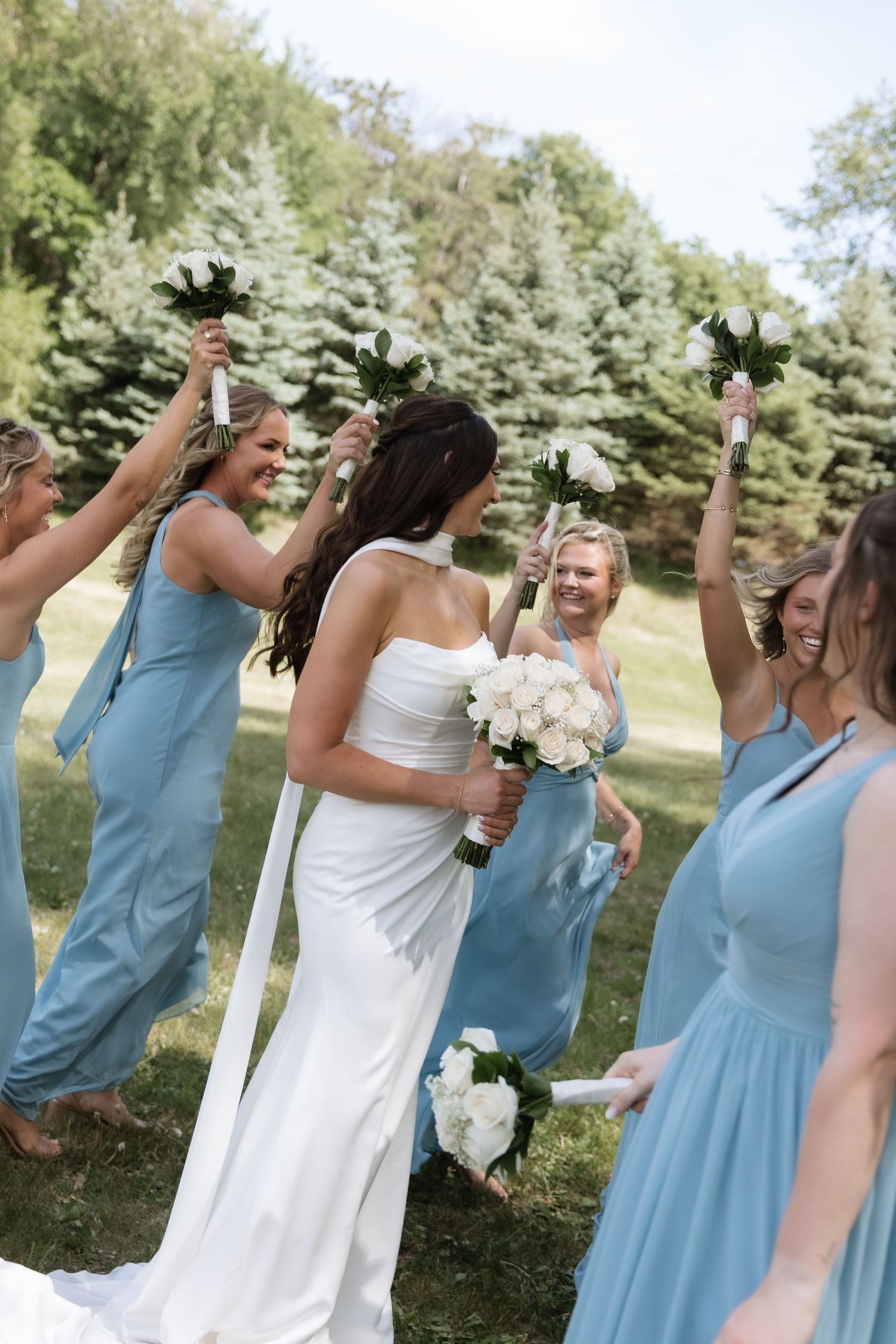 Bride in white gown surrounded by bridesmaids in blue dresses raising white bouquets outdoors.