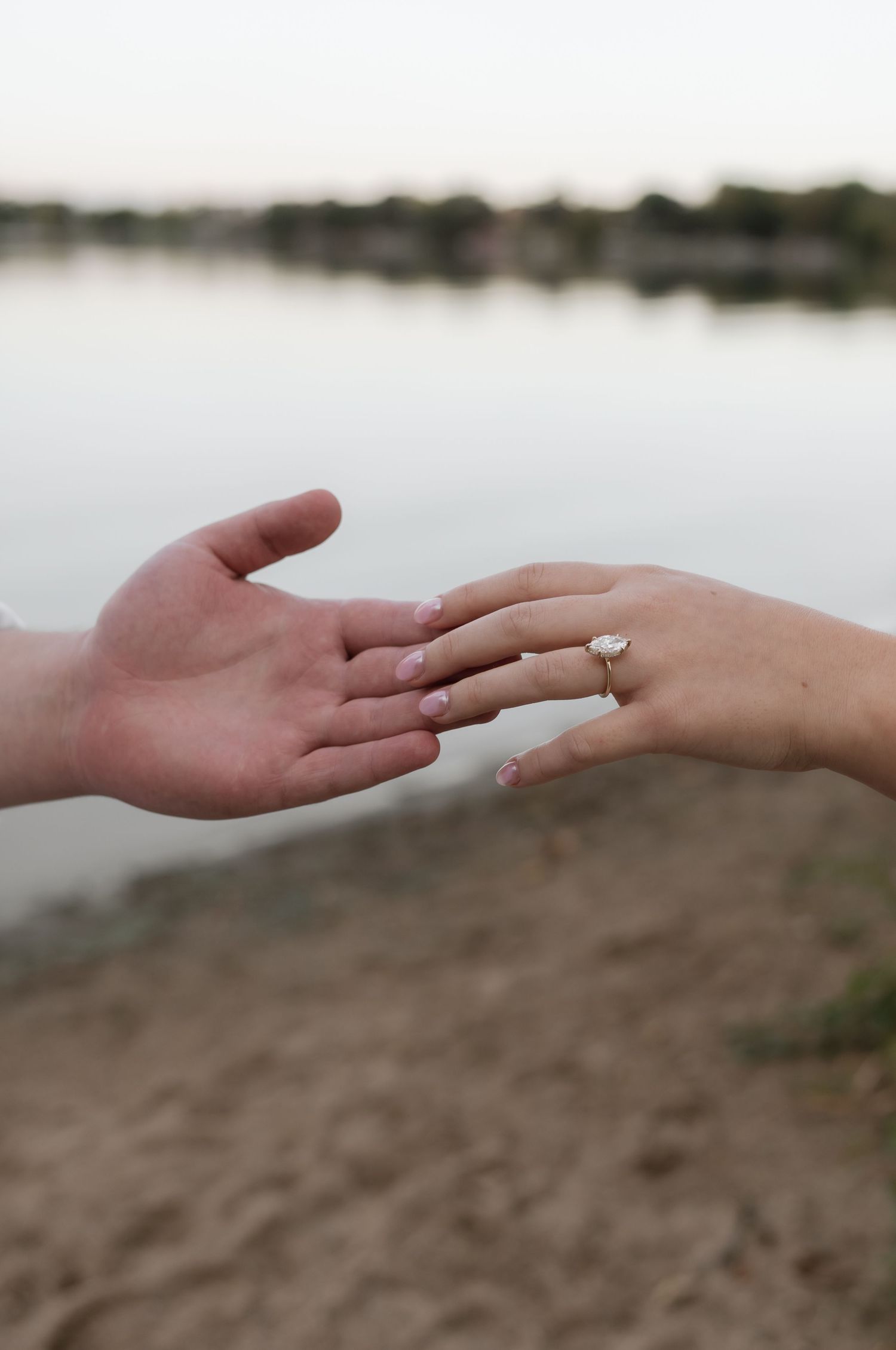 Two hands reaching toward each other near a lake, with a diamond engagement ring on the woman's finger.