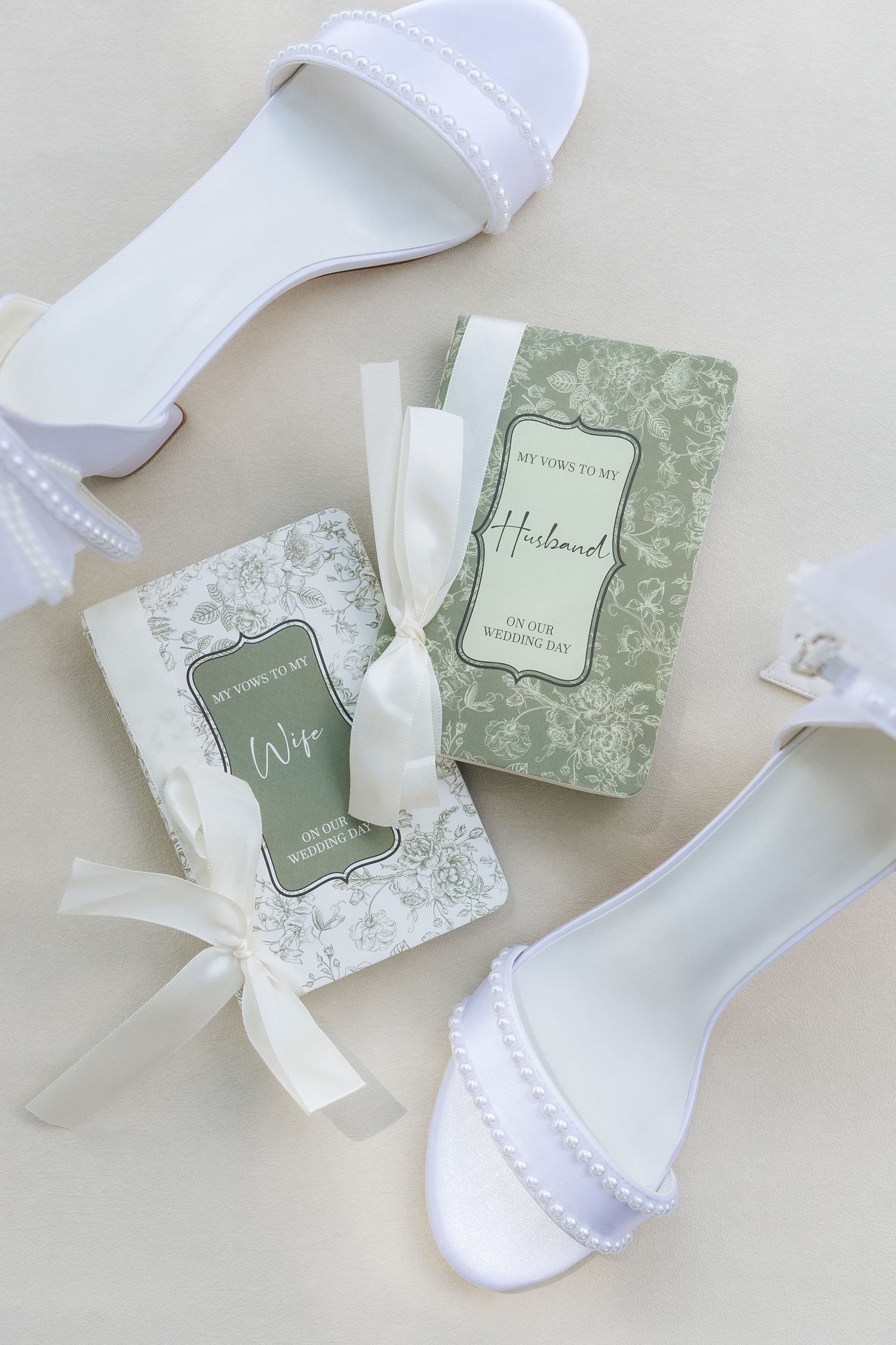 White bridal sandals with pearl details beside sage green and white vow books tied with ivory ribbon on a neutral background.