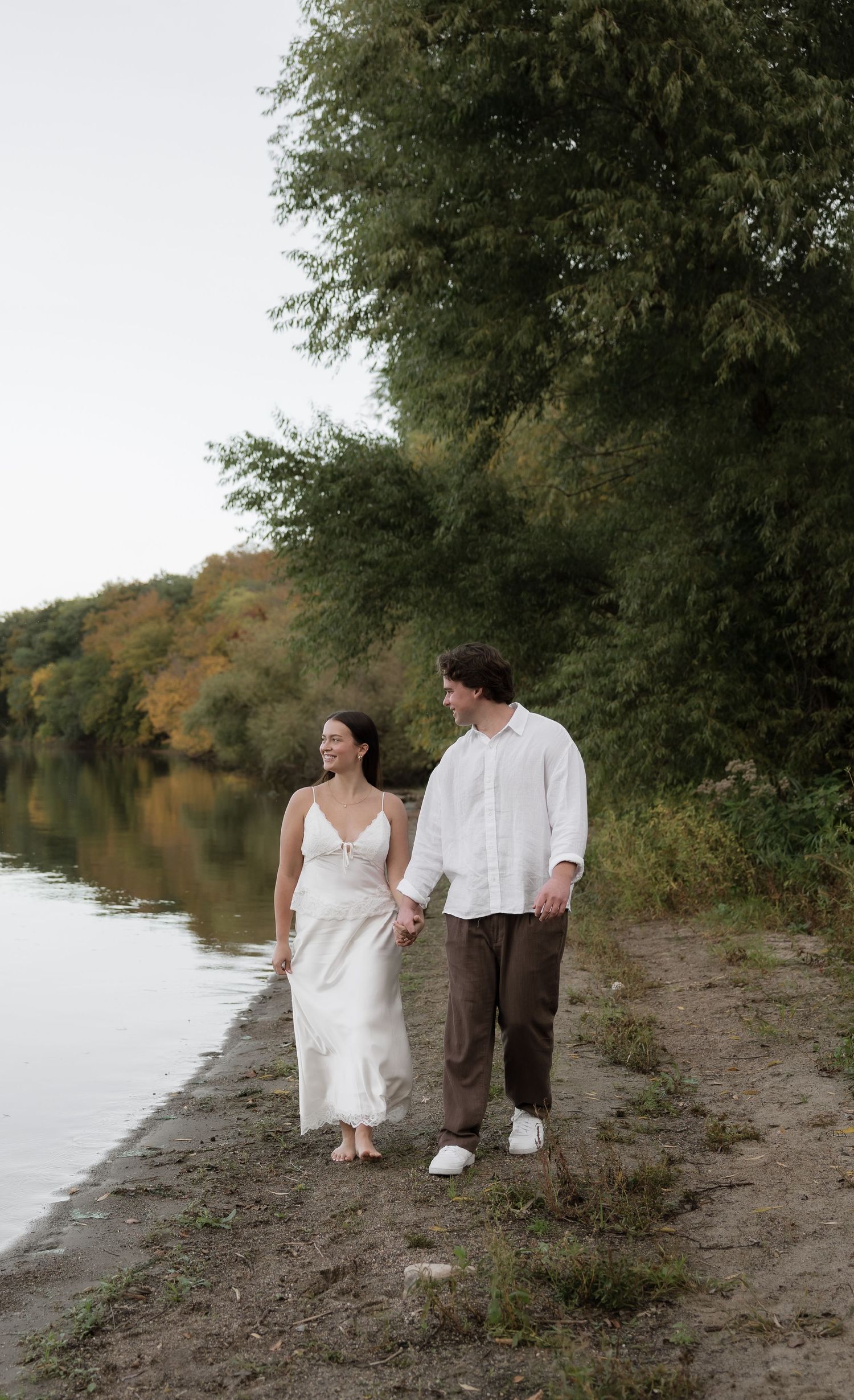 A couple walks hand-in-hand along a serene riverside path surrounded by lush green and autumn trees.