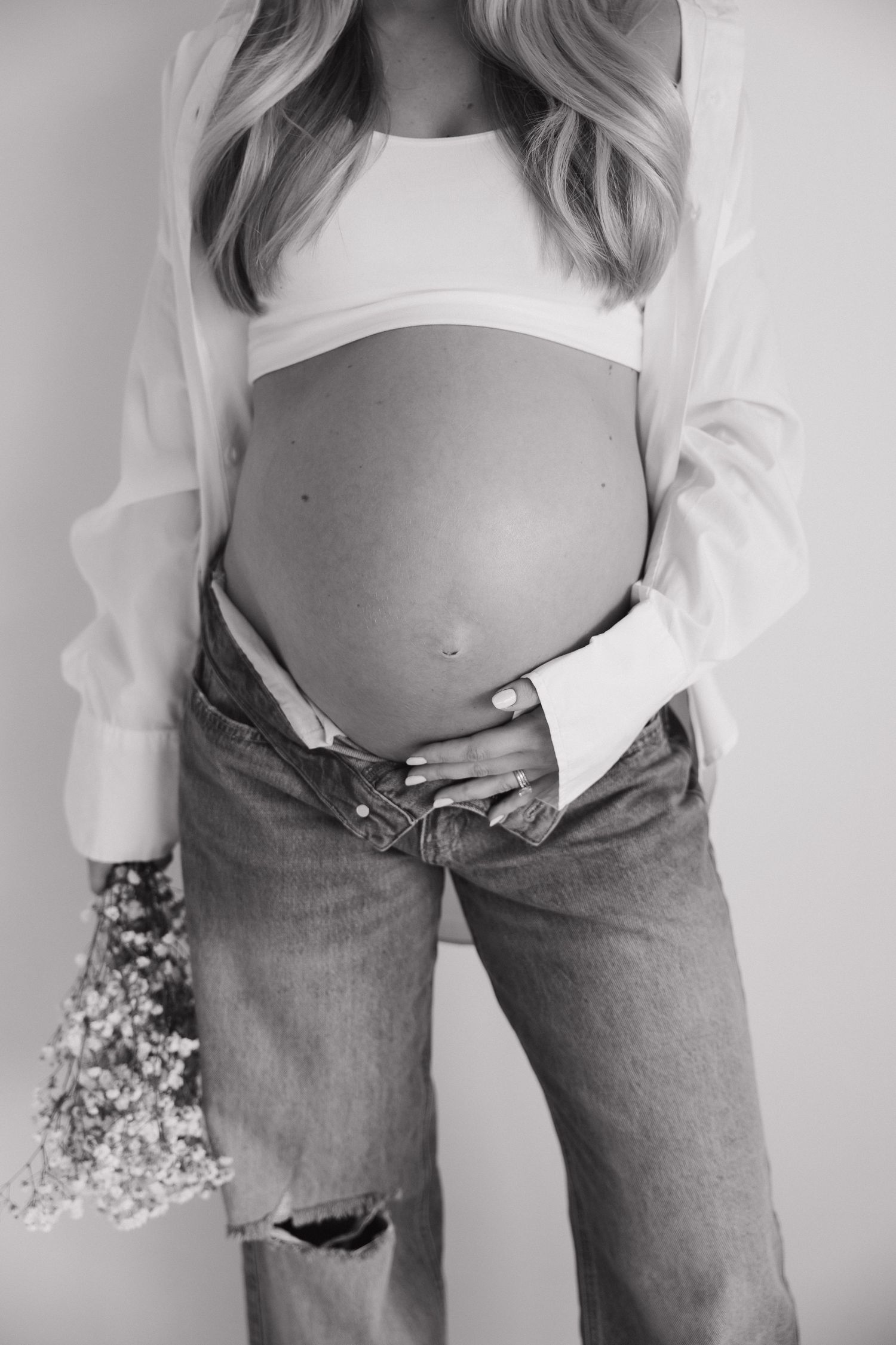 Black and white photo of pregnant woman holding baby bump in white crop top and ripped jeans with flowers.