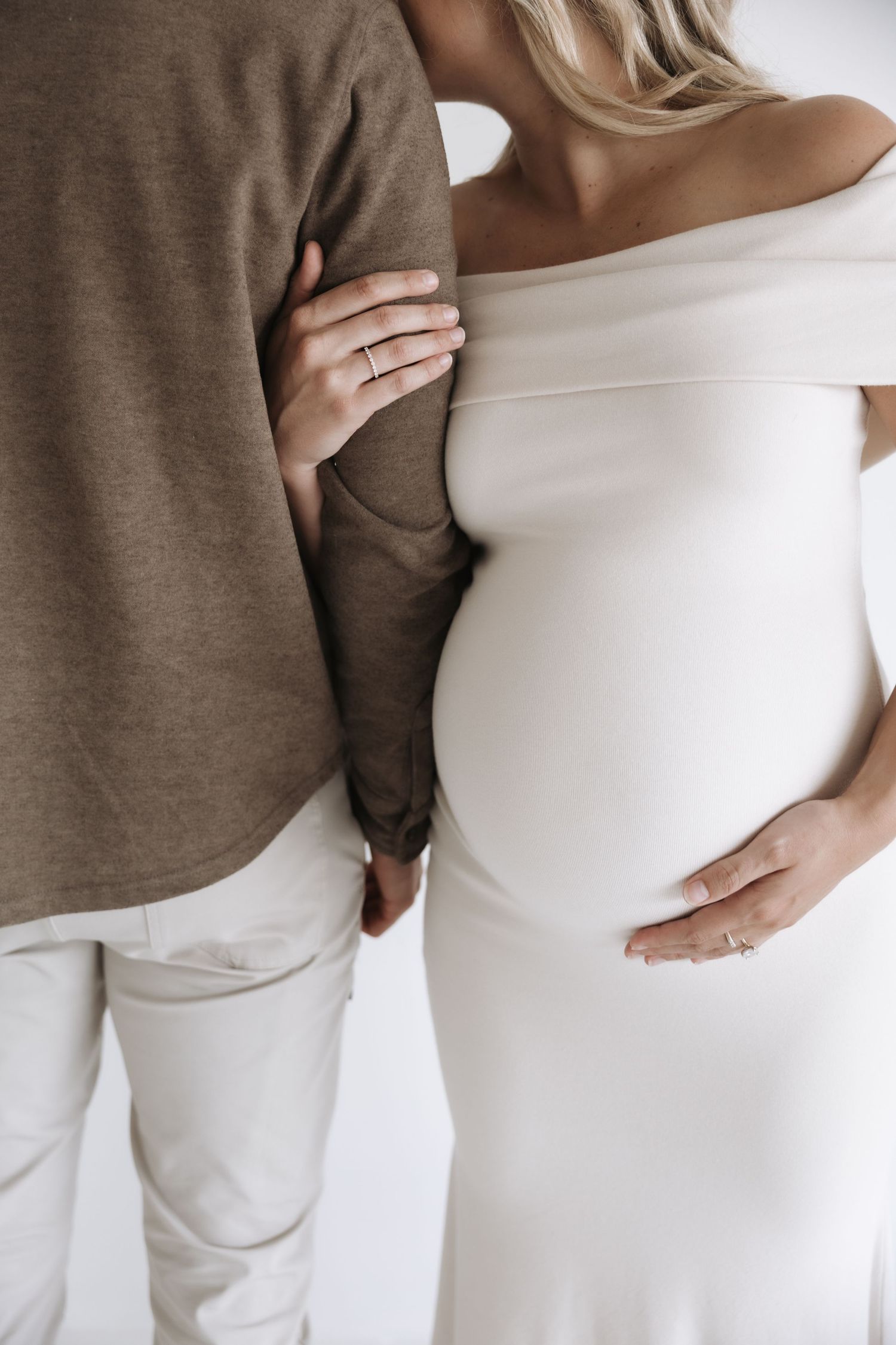 Pregnant woman in white off-shoulder dress embracing partner from behind during maternity session.