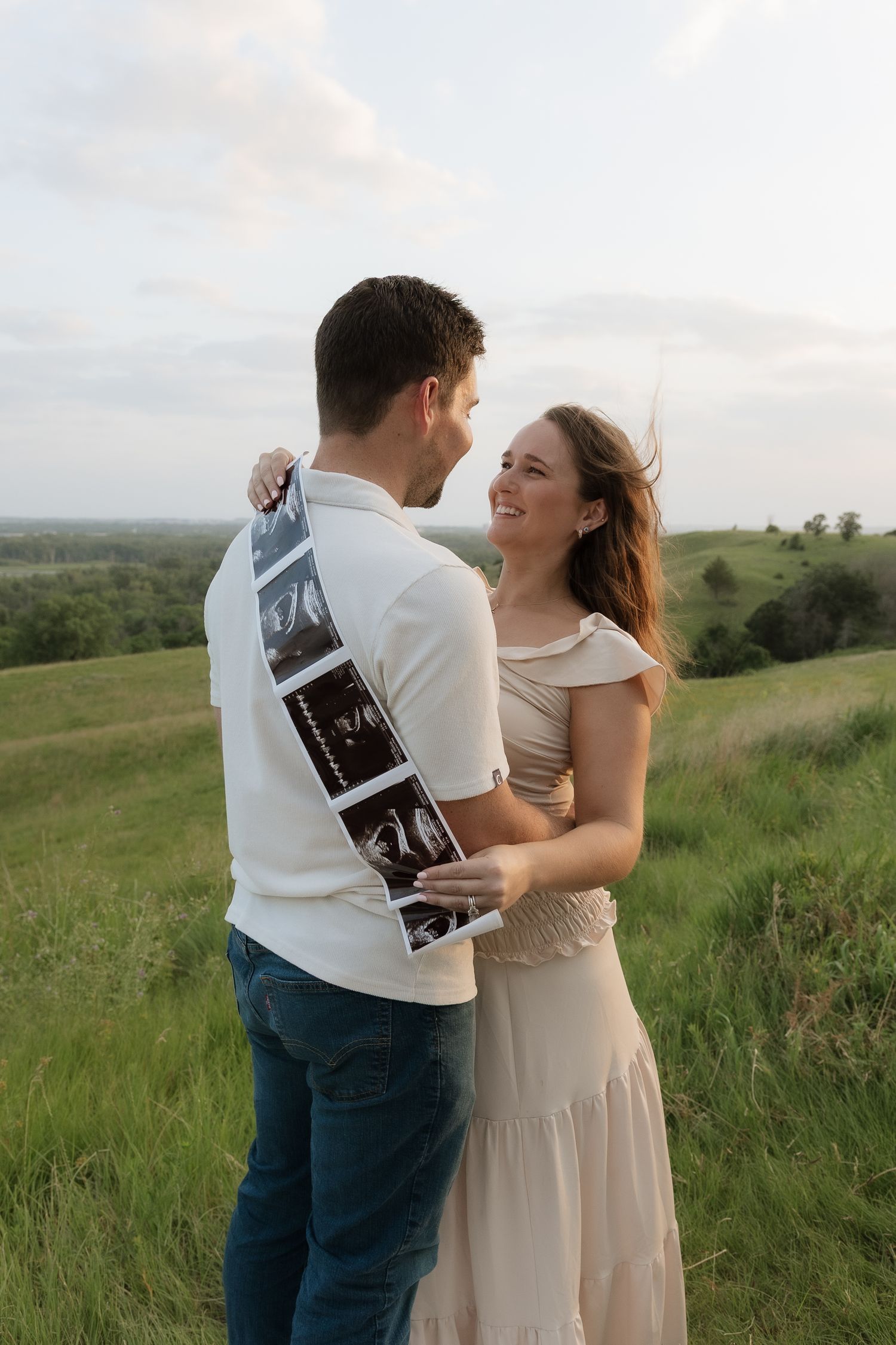 Happy couple embracing outdoors holding ultrasound photos, announcing pregnancy in a lush green field.