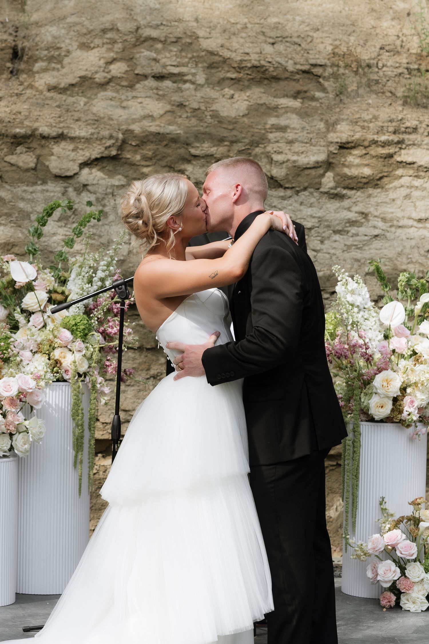 Bride and groom share first kiss at wedding altar surrounded by blush floral arrangements against stone wall.
