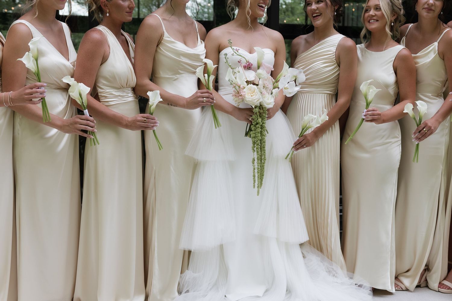 Bride with cascading bouquet poses with bridesmaids in matching cream one-shoulder gowns outdoors.