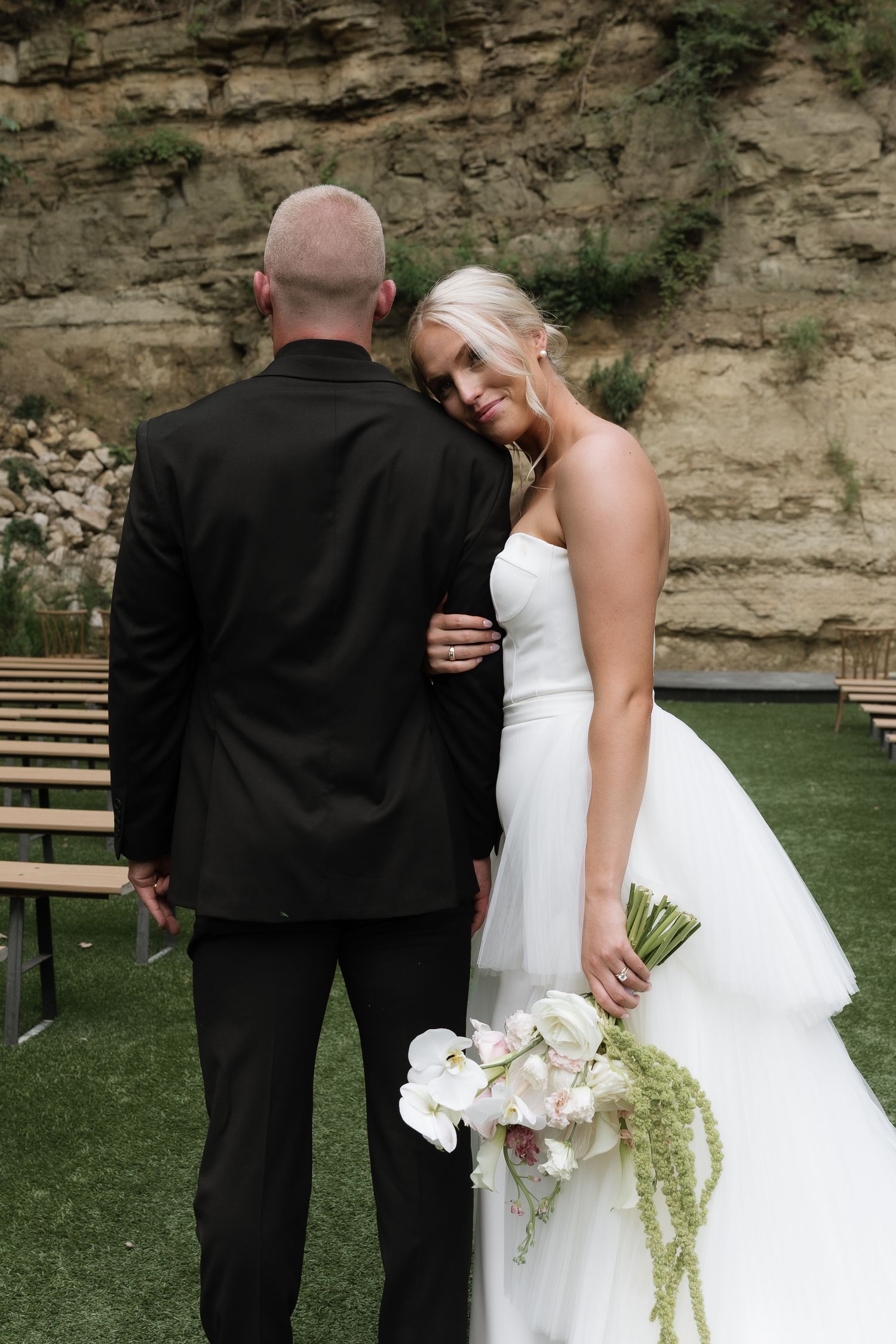 Bride in white strapless gown holds floral bouquet, smiling at groom in black suit at outdoor wedding venue.