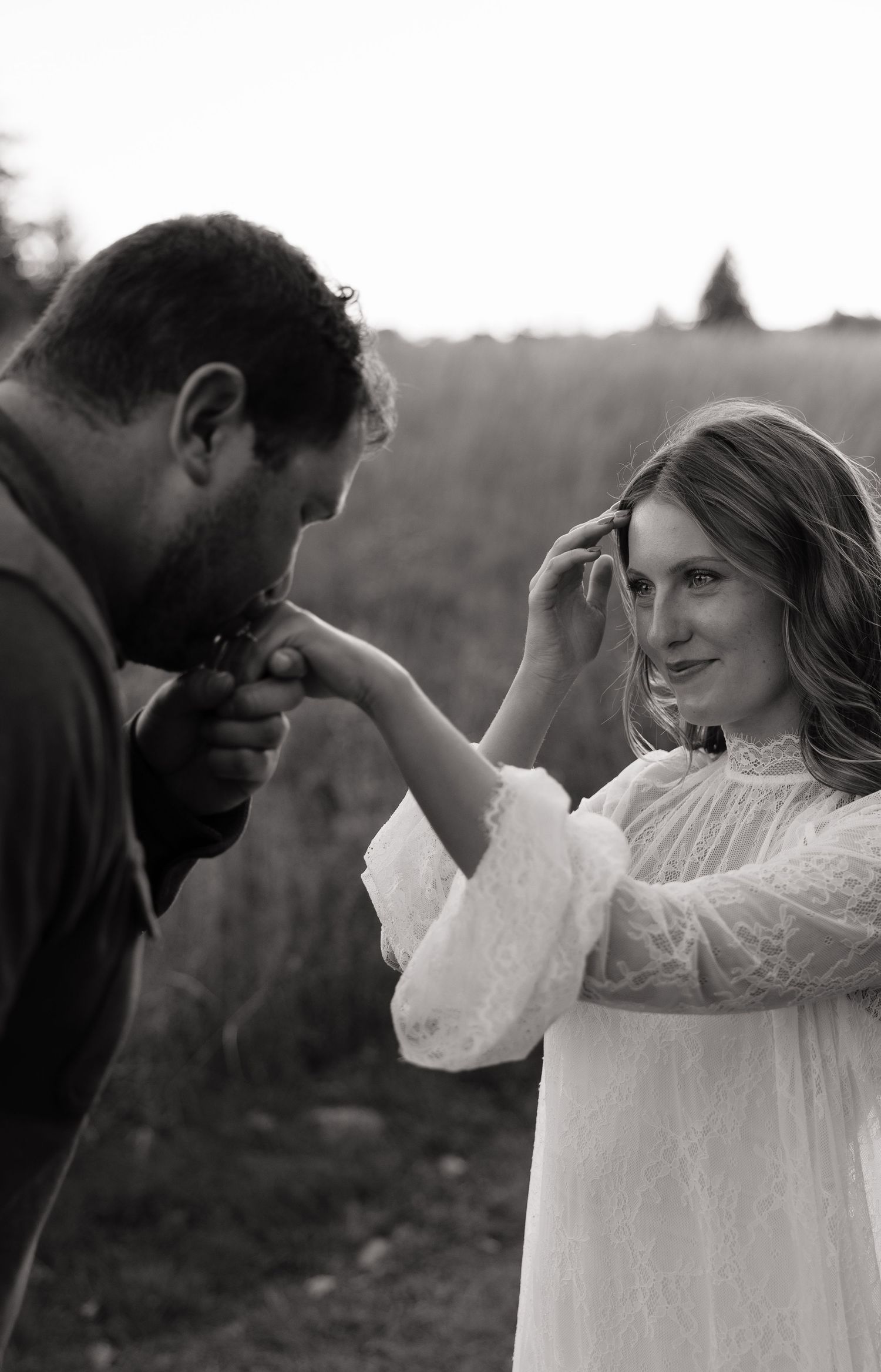 A groom kisses the hand of his smiling bride in a lace dress during an outdoor wedding ceremony.