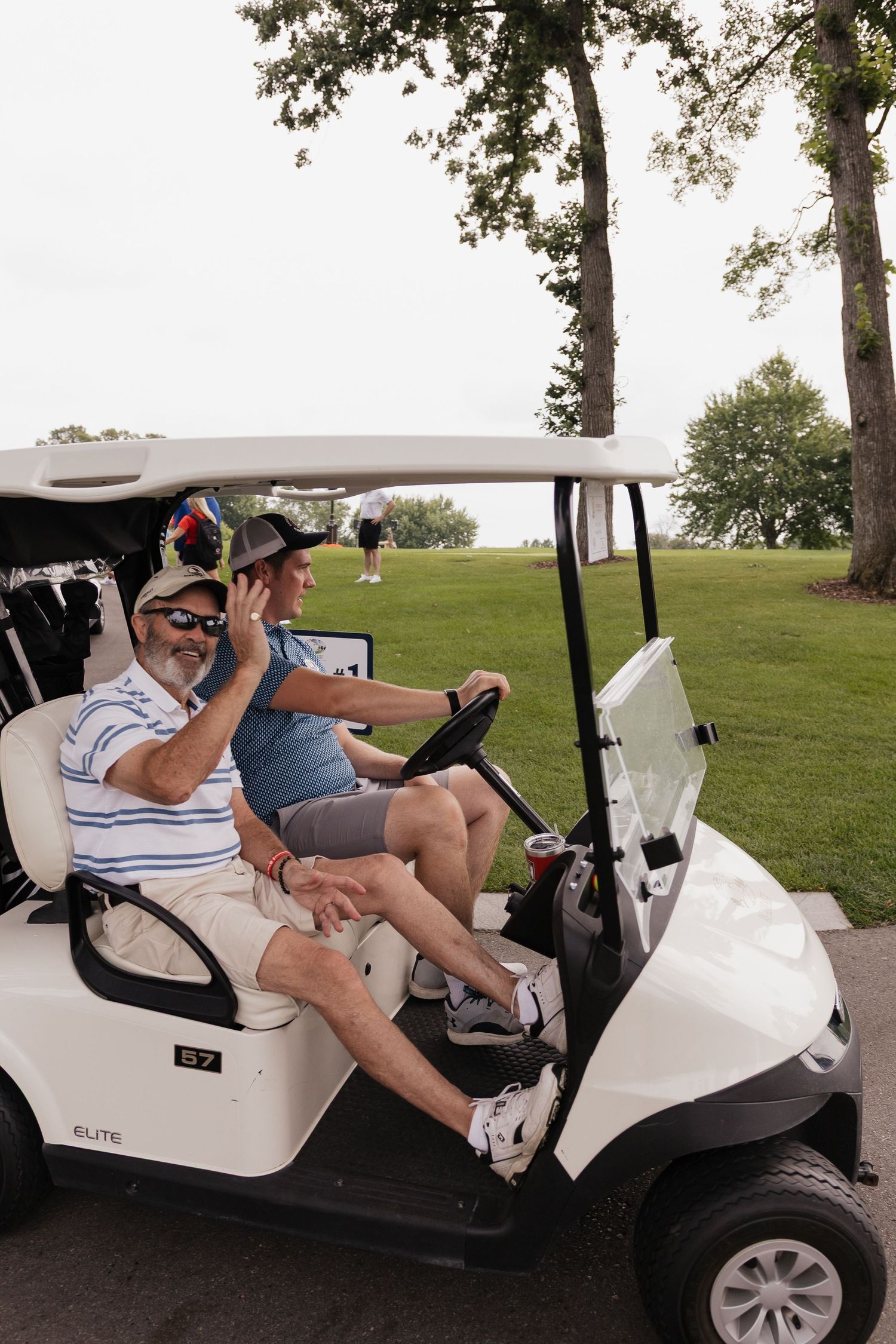 Two men riding in a white Elite golf cart on a sunny golf course, one waving at the camera.