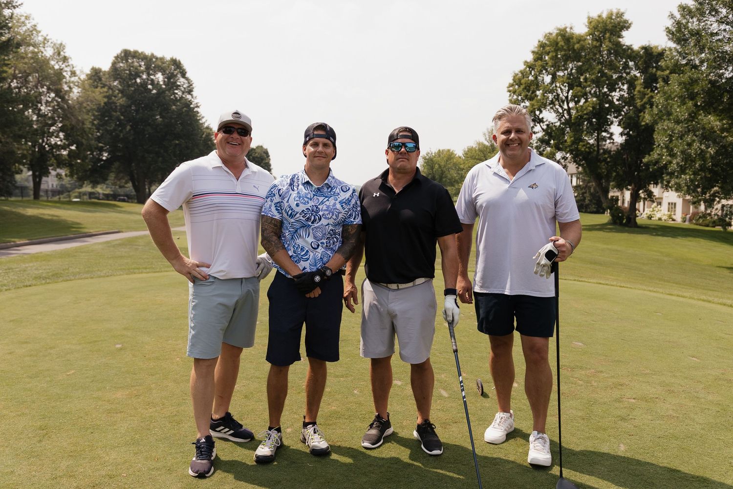 Four men posing together on a golf course on a sunny day, wearing casual golf attire.