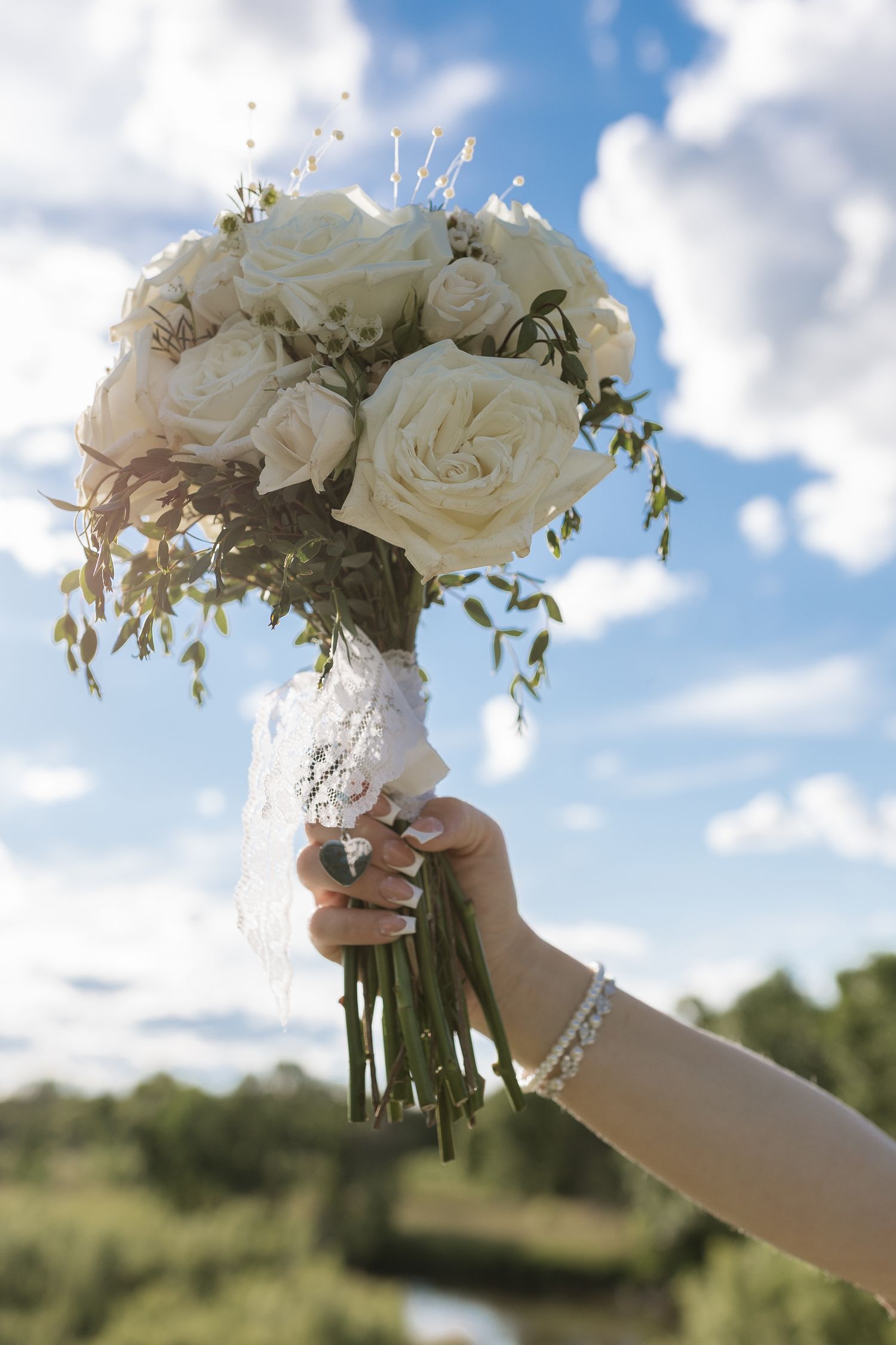 Bride holding white rose wedding bouquet with greenery and lace ribbon against a blue sky backdrop.
