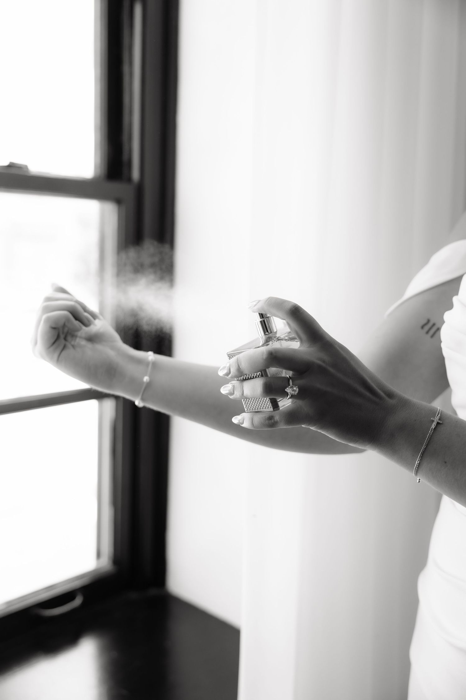 Black and white photo of a bride spraying perfume on her wrist near a window on her wedding day.