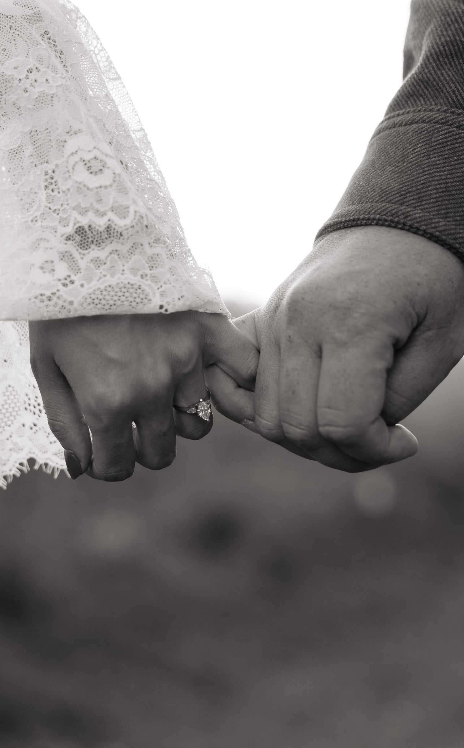 Black and white photo of a couple holding hands, woman wearing an engagement ring, dressed in lace wedding attire.