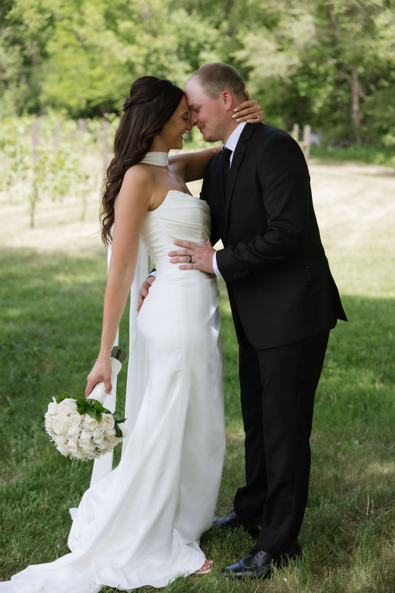 Bride and groom share a romantic almost-kiss, bride holding white rose bouquet in a garden setting.