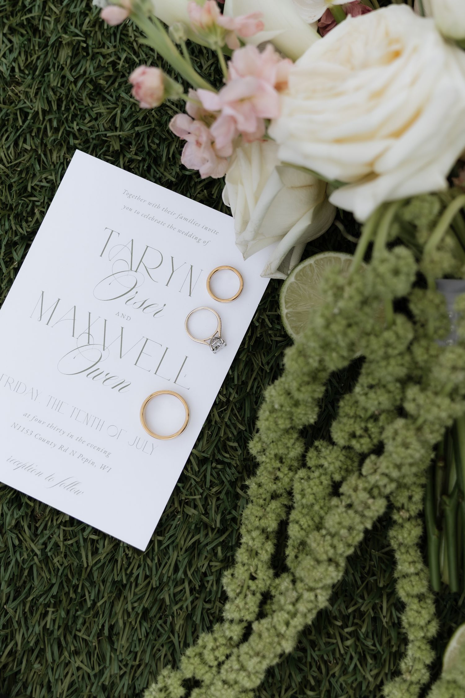 Wedding invitation with gold rings and diamond engagement ring on green grass beside white roses and pink floral bouquet.