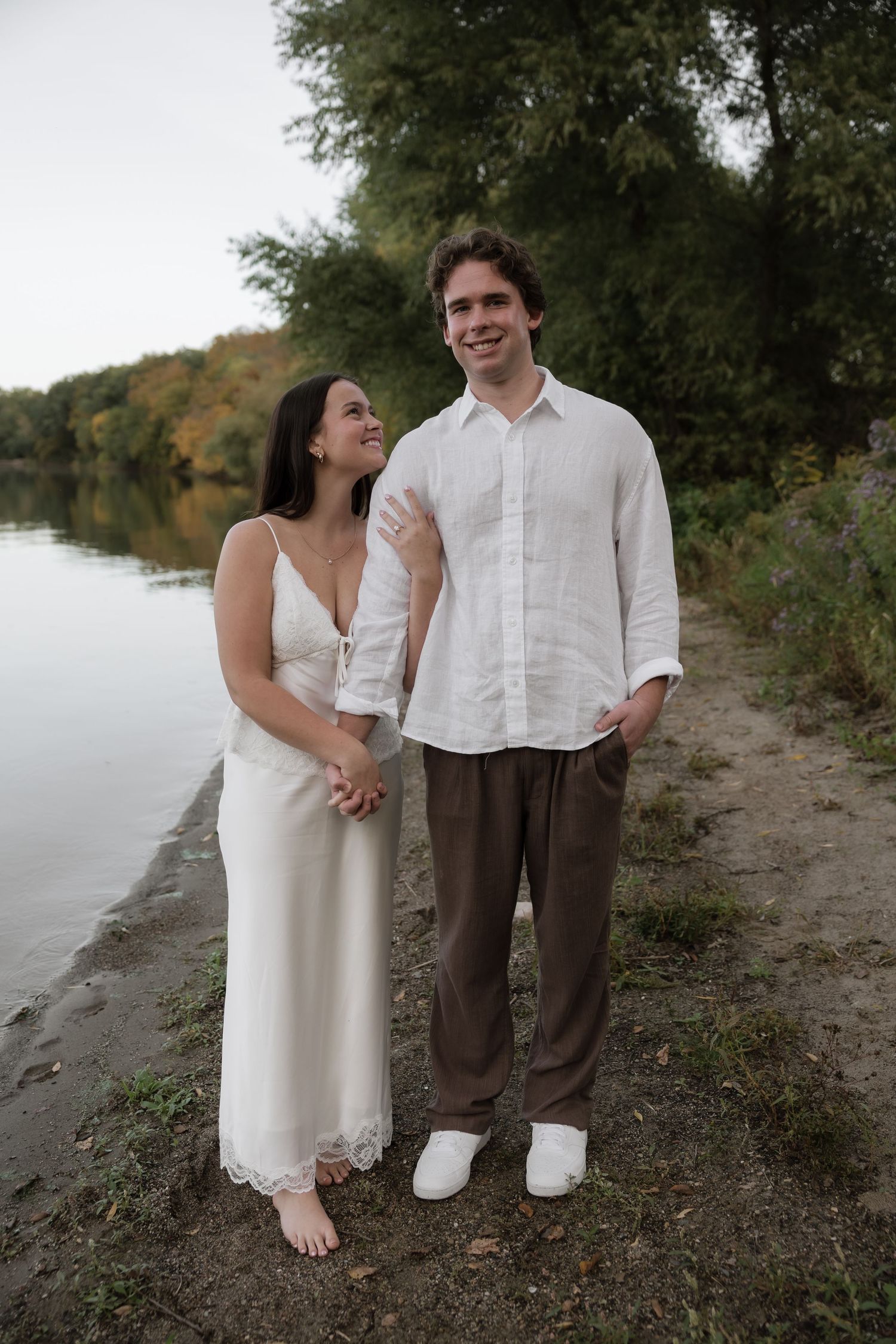 Engaged couple holding hands by a lake, woman in white lace dress and man in white linen shirt smiling.