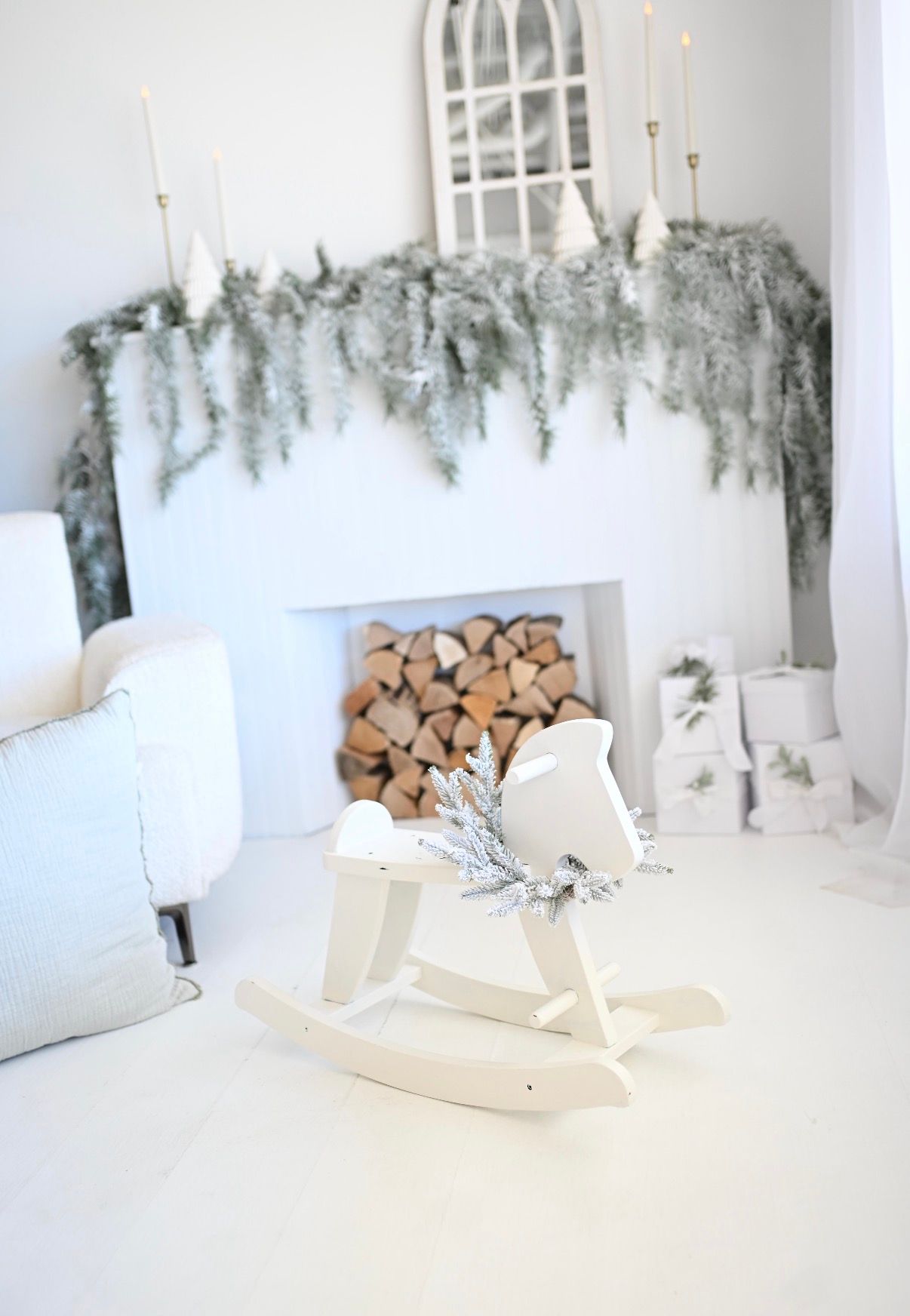 White rocking horse toy in front of a festive fireplace with snowy garland, candles, and stacked firewood.