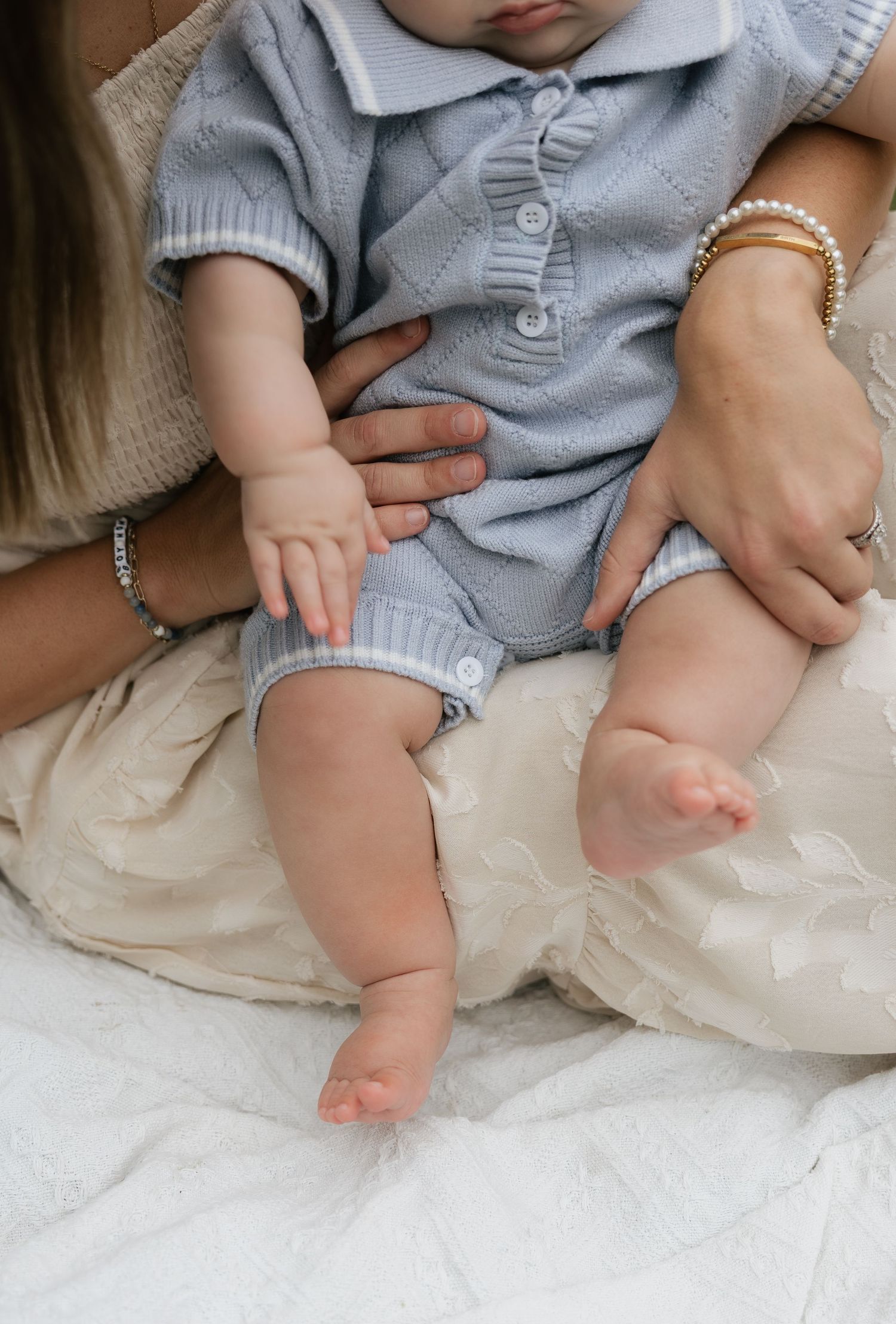 Baby in blue knit polo onesie held by mother wearing pearl bracelet, sitting on white fabric.