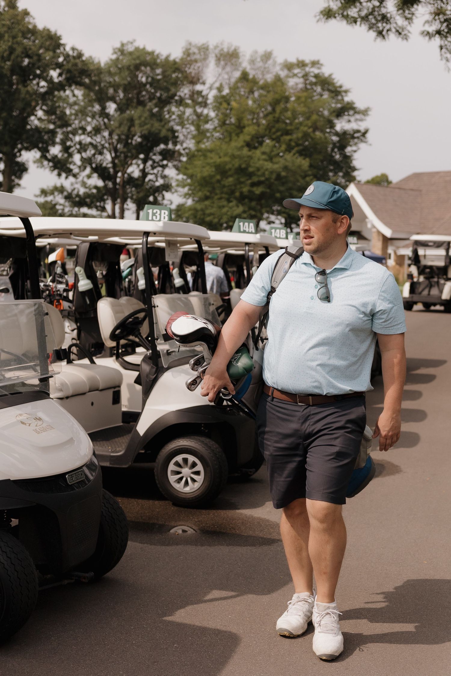 Man in blue polo and cap carrying golf bag walking past row of golf carts at a golf course.