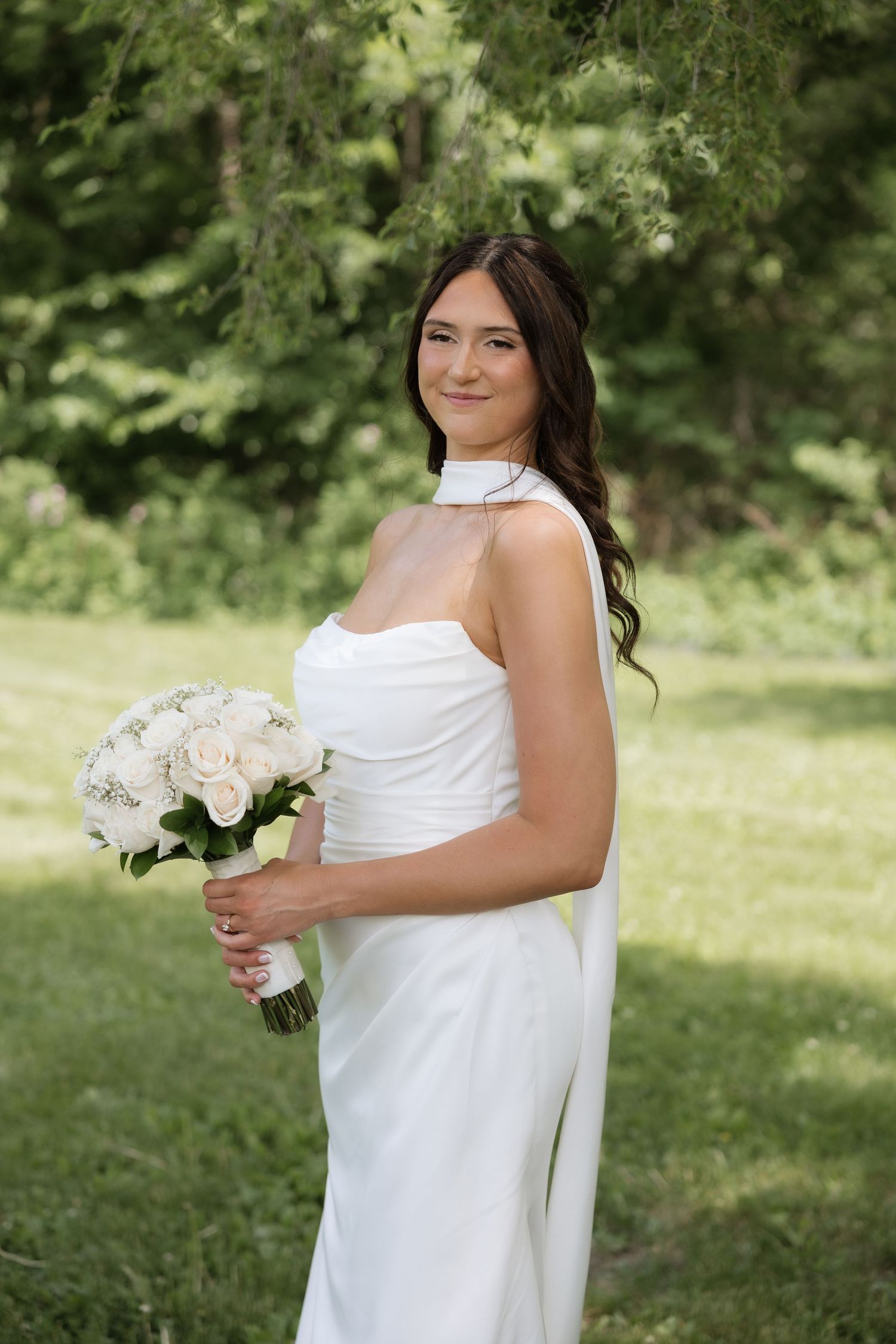 Smiling bride in a white halter wedding dress holds a bouquet of white roses in a lush green outdoor setting.