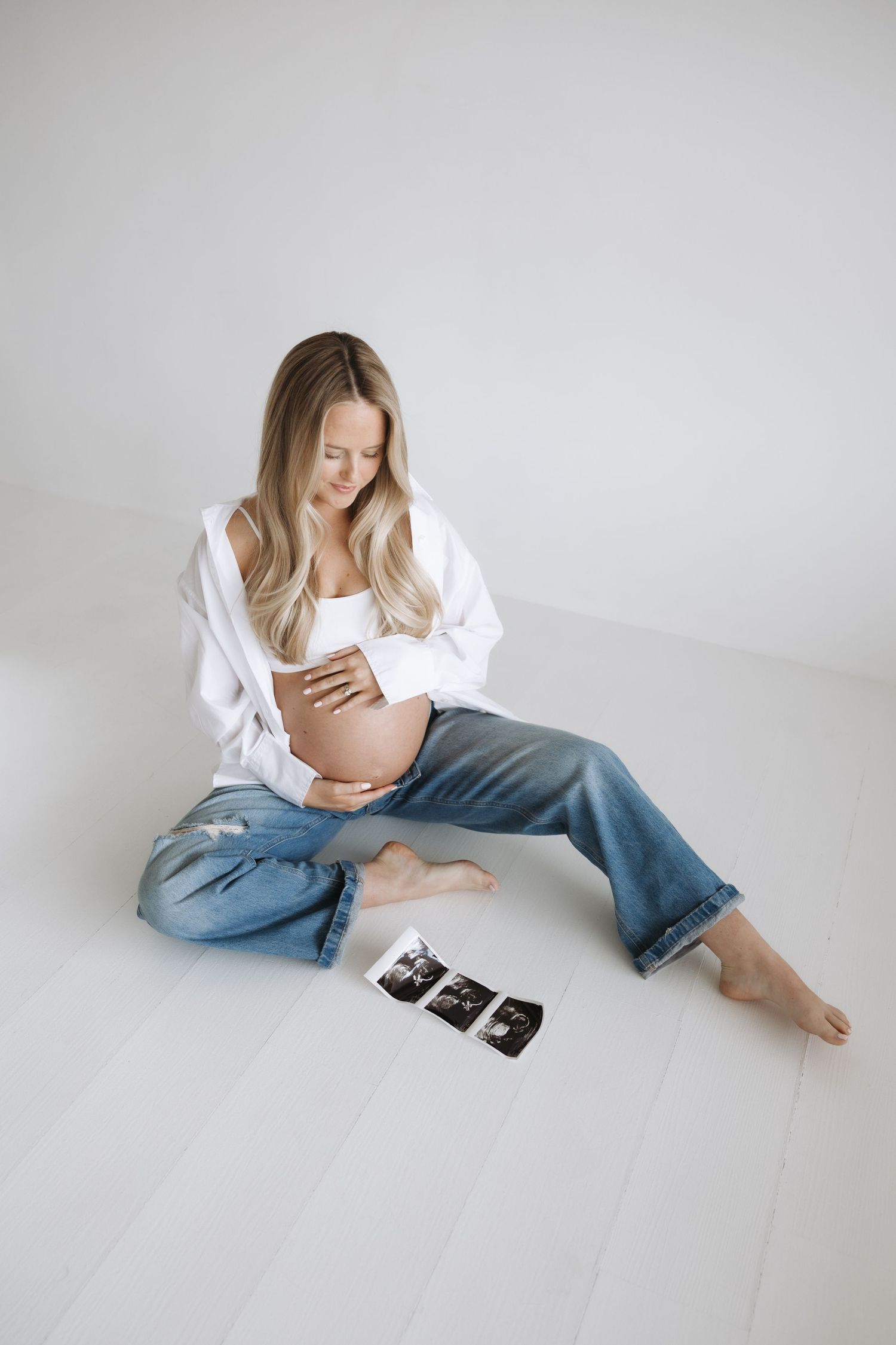 Pregnant woman in white shirt and jeans sitting on floor smiling at baby bump with ultrasound photos nearby.