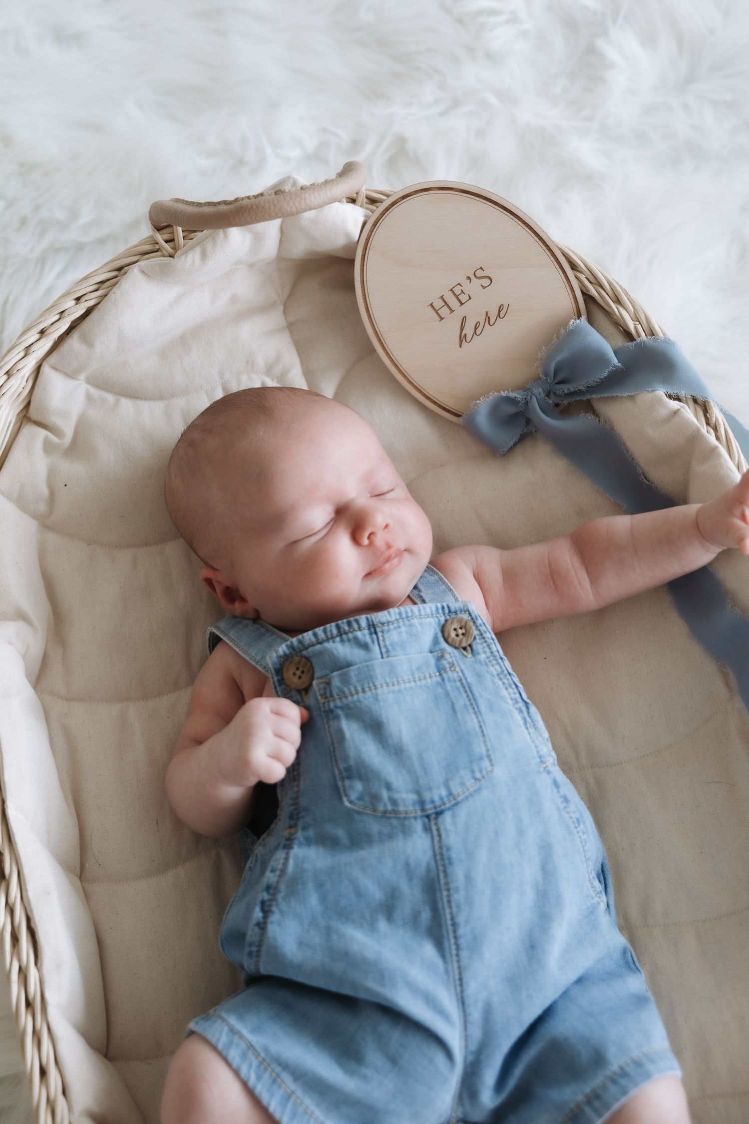 Sleeping newborn baby in denim overalls beside a wooden he's here announcement sign in wicker basket.