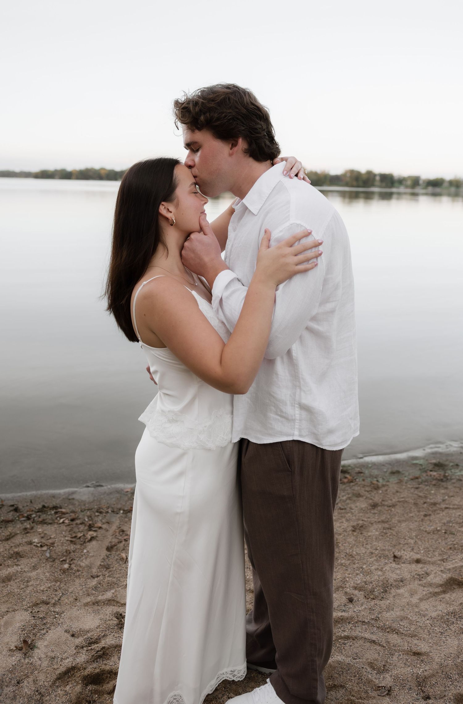 Couple sharing a romantic kiss on a sandy beach shore, woman in white dress, man in white shirt and brown pants.