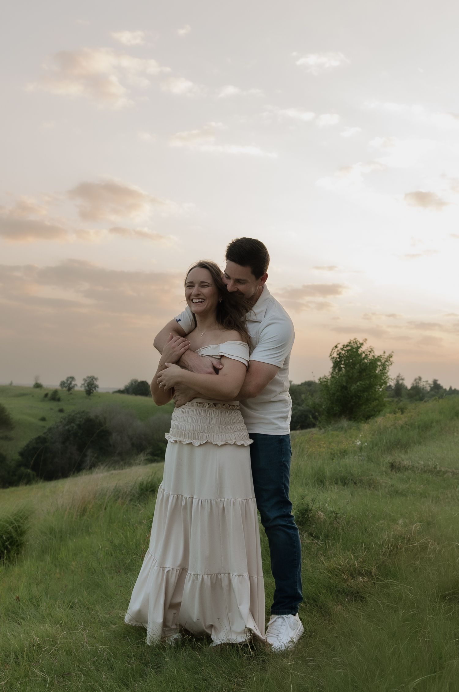 Couple embracing on green hillside at sunset, woman in white dress smiling as man hugs her from behind.